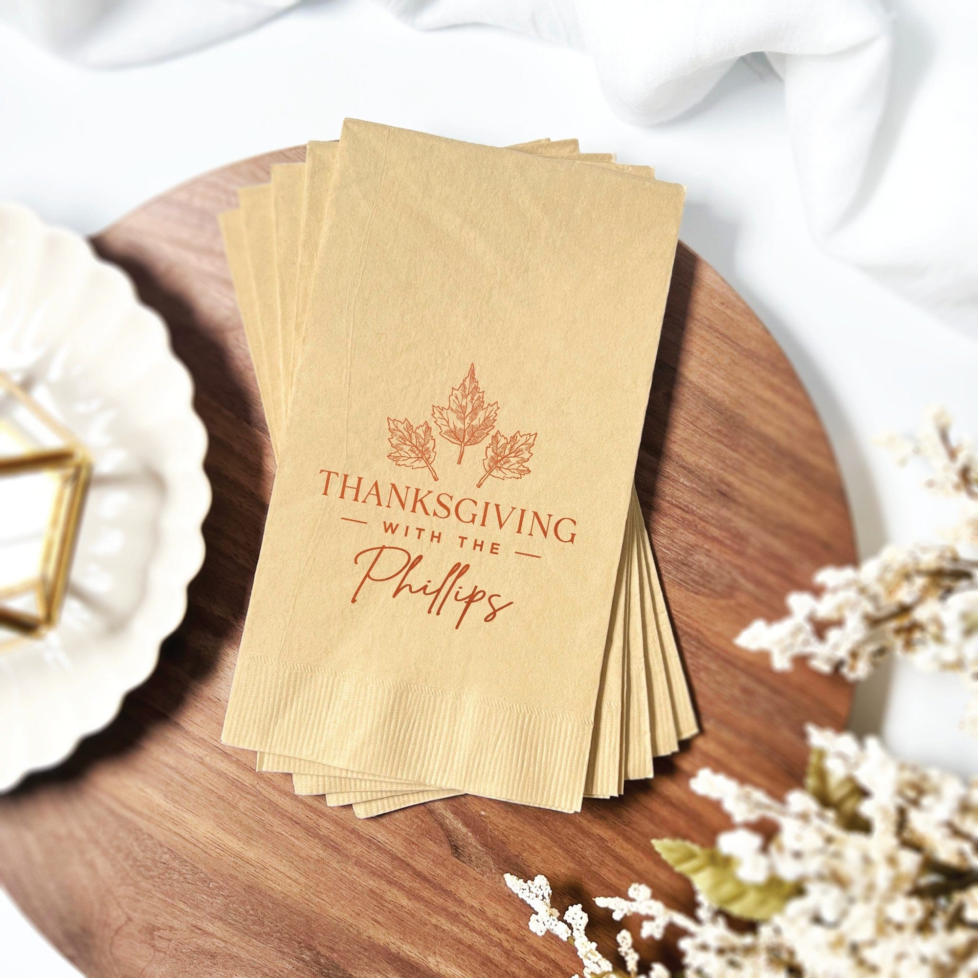 A stack of rectangular kraft-colored paper guest towels is displayed on a wooden board, personalized with a reddish-brown maple leaf design for a festive Thanksgiving celebration.