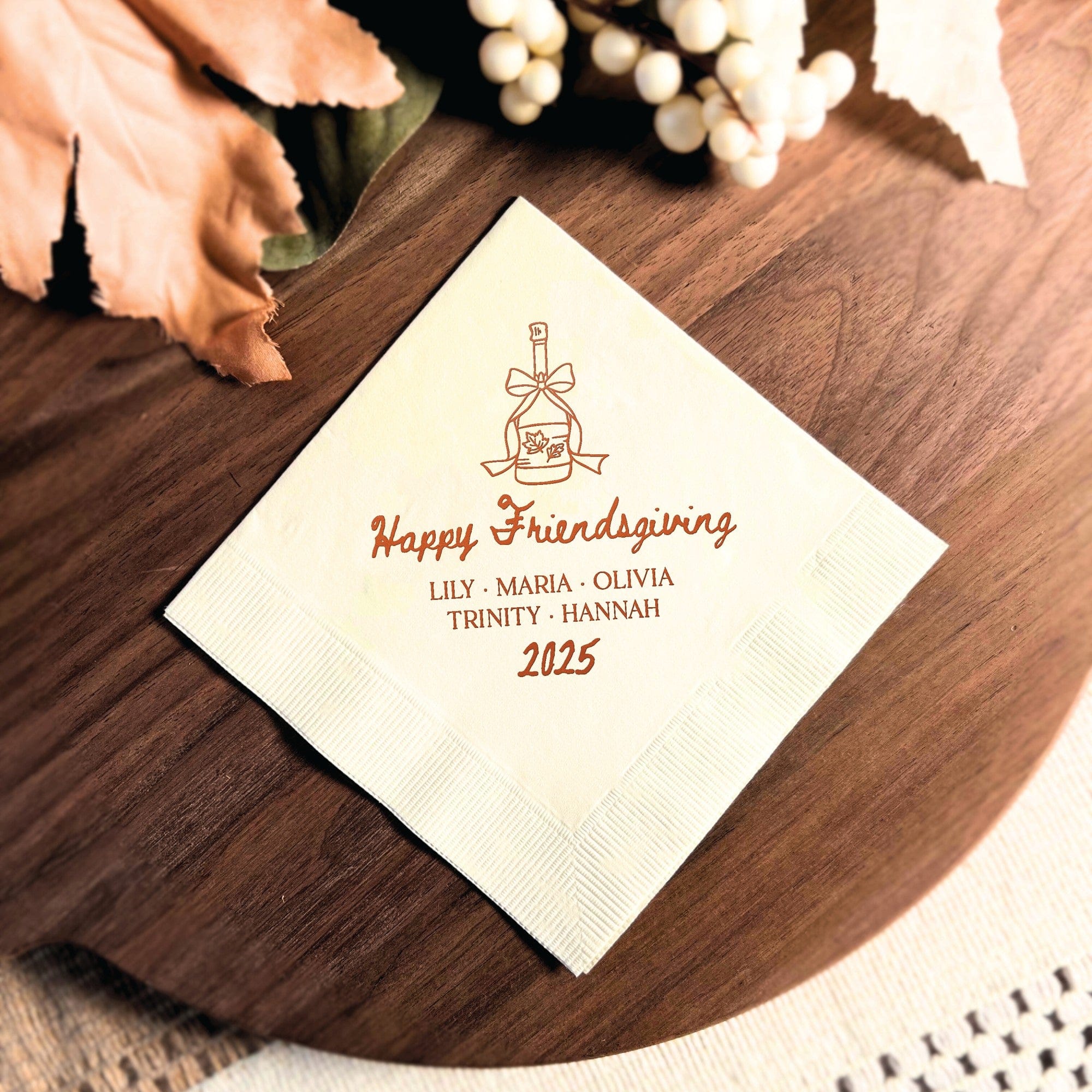 A cream-colored paper napkin featuring a custom reddish-brown print of a decorative bottle with fall leaves, suitable for a Friendsgiving or autumn celebration.