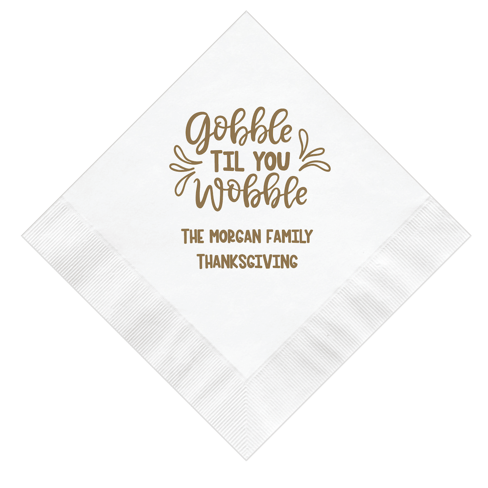 White napkin with gold foil stamped text reading "Gobble Til You Wobble" and "The Morgan Family Thanksgiving"