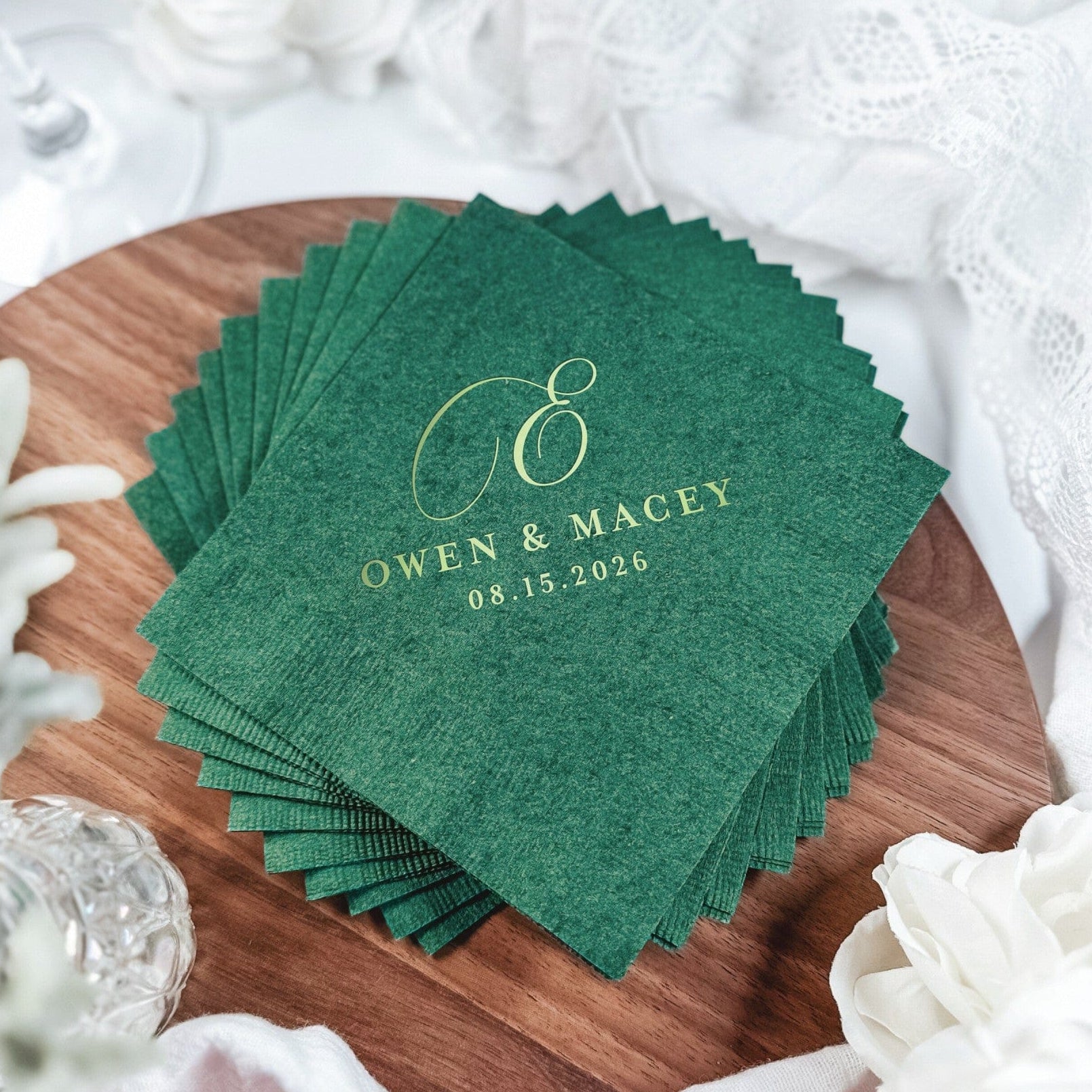 A stack of square, dark green paper wedding napkins featuring a custom gold foil initial and text, displayed on a wooden surface with white floral decor and lace, perfect for personalized event tables.