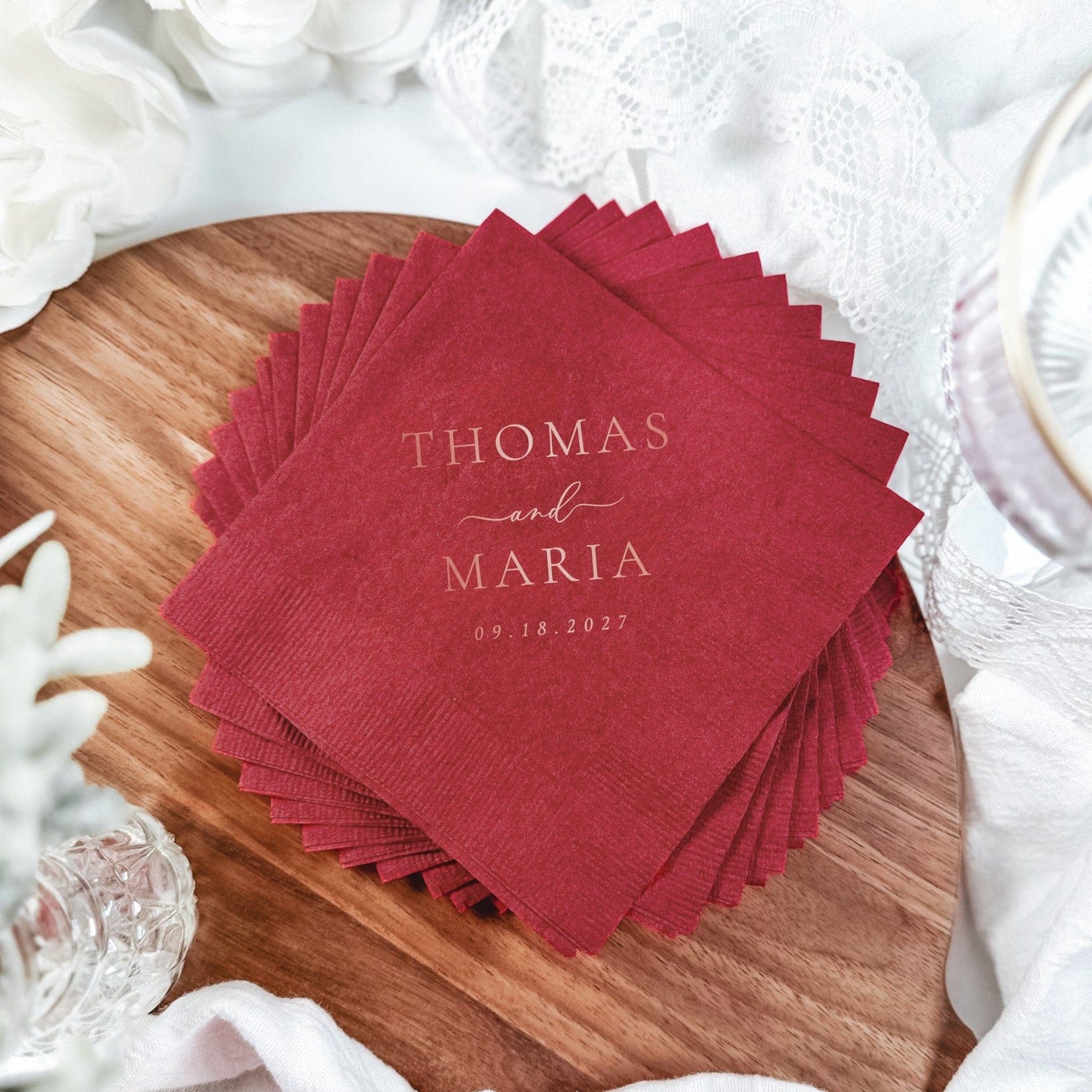 A stack of textured red disposable paper napkins is prominently displayed on a wooden surface, with the top napkin featuring elegant, personalized foil-printed text, perfect for a wedding or special event.