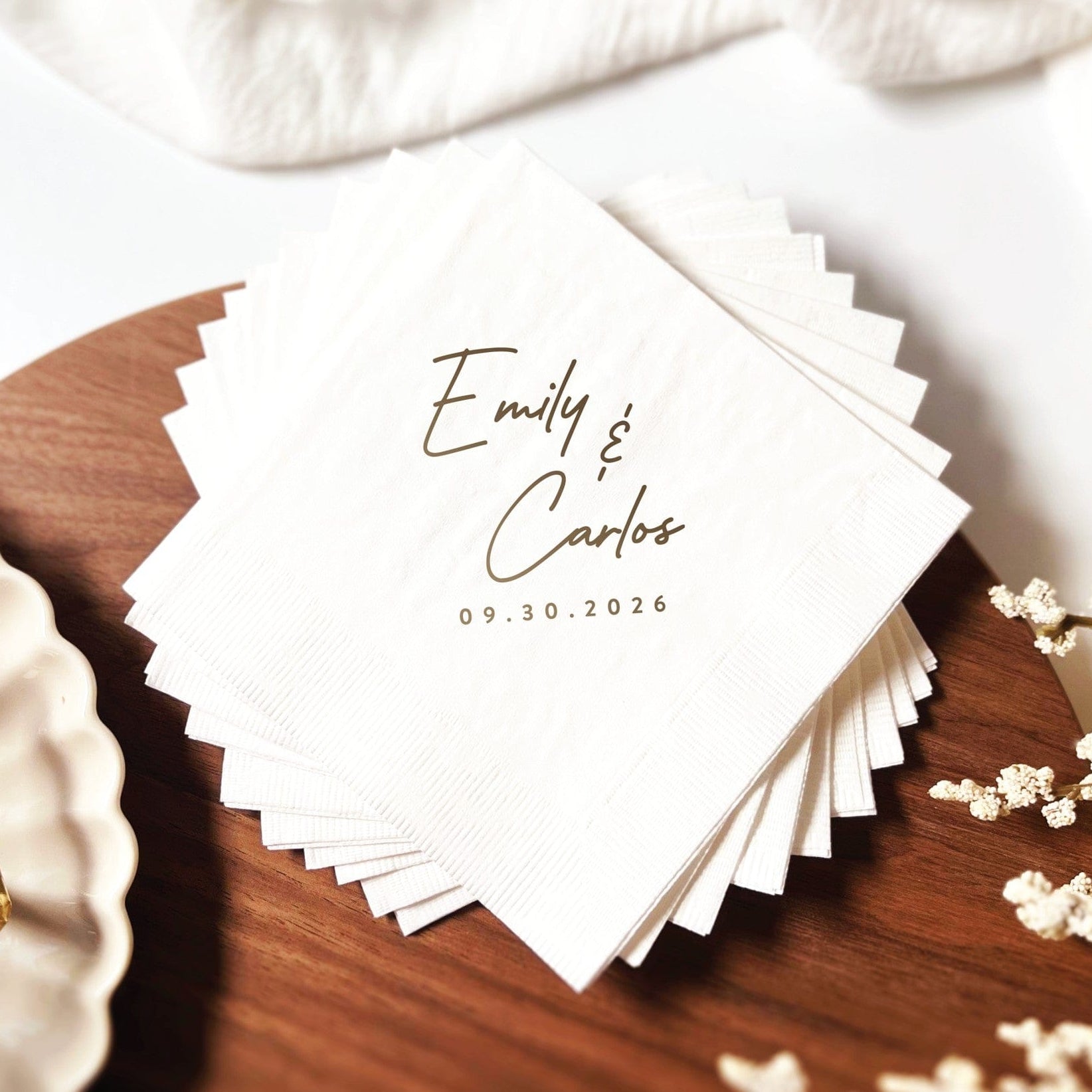 A stack of personalized white paper wedding napkins featuring elegant custom script text, fanned out on a dark wooden surface.