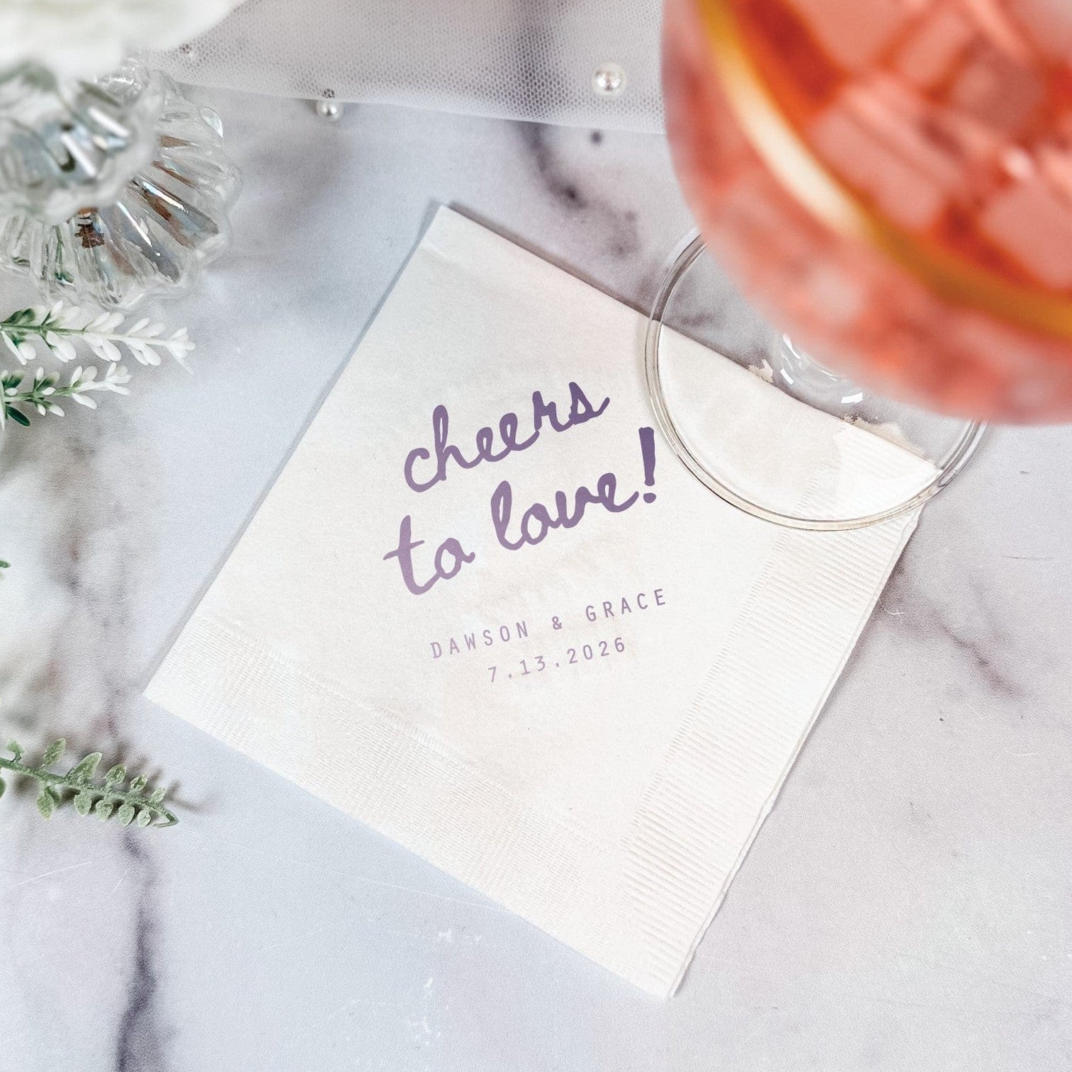 A white paper wedding napkin with personalized purple text and a border texture, displayed on a marble background with a drink and floral accents.