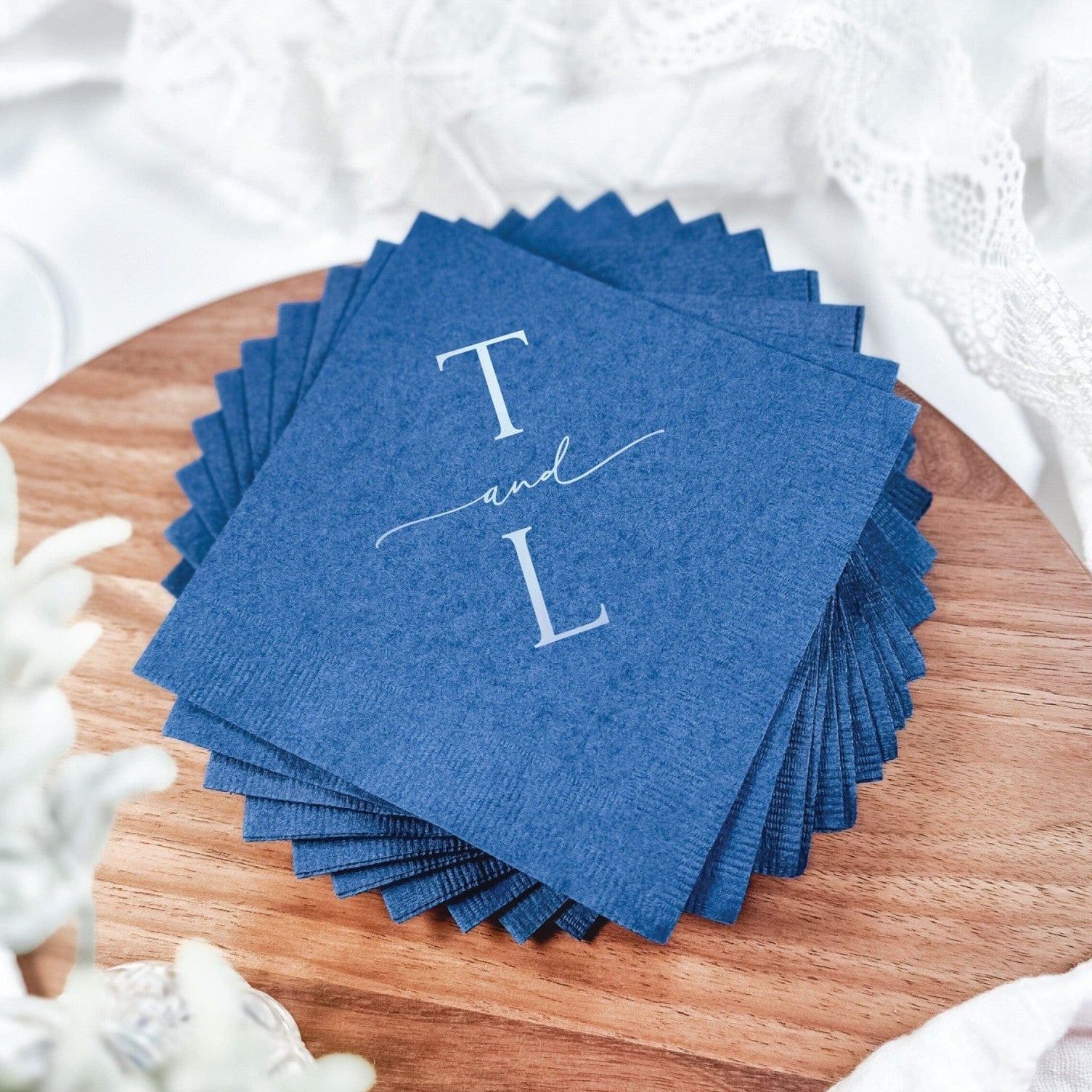 A stack of elegant deep blue paper napkins featuring custom white printing, arranged on a wooden board for a wedding reception or special event table setting.