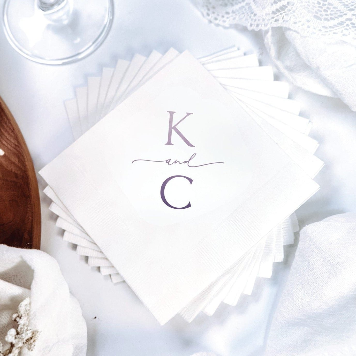 A stack of white paper wedding napkins featuring personalized initials and an 'and' in purple print, designed for elegant event decor.
