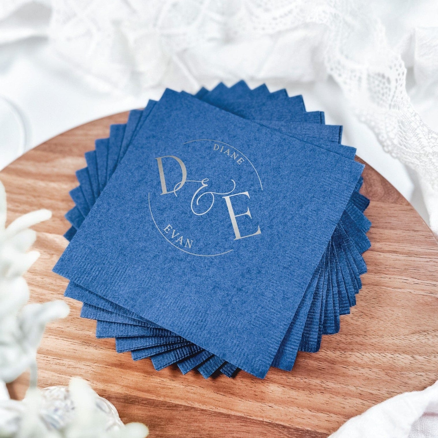A stack of deep blue paper cocktail napkins featuring a circular metallic foil monogram design, perfect for a wedding or special event.