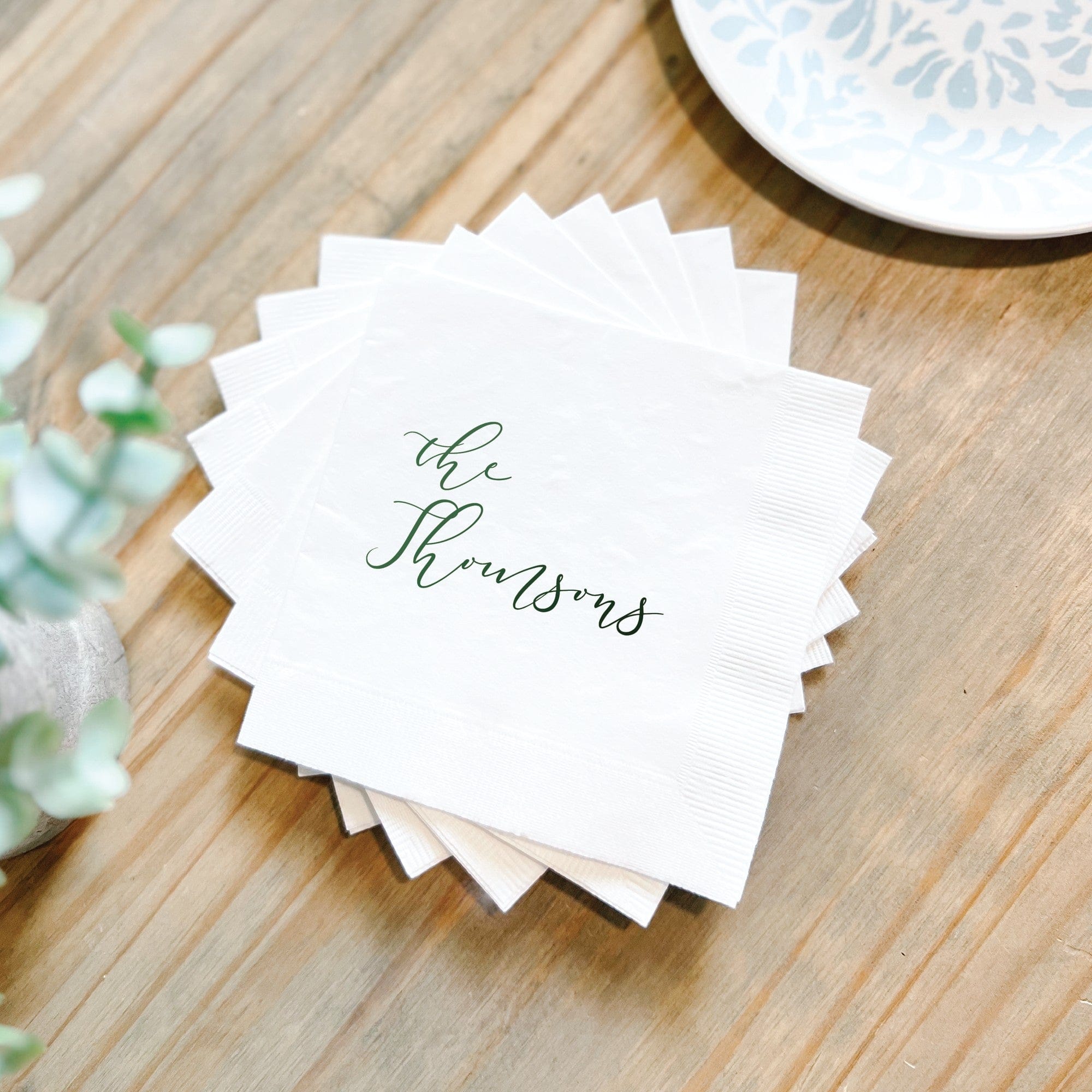 A stack of white paper wedding napkins personalized with custom script lettering in emerald green, suitable for a wedding or special event.