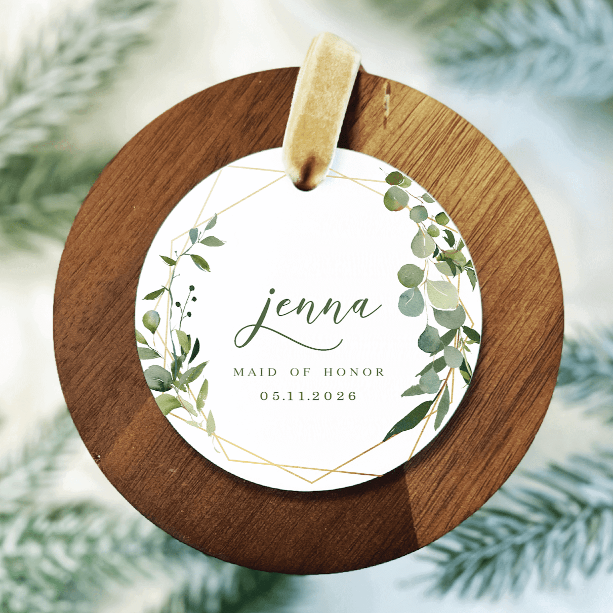 Round wooden ornament with white acrylic center featuring "jenna," "maid of honor," and date "05.11.2026" surrounded by green foliage design