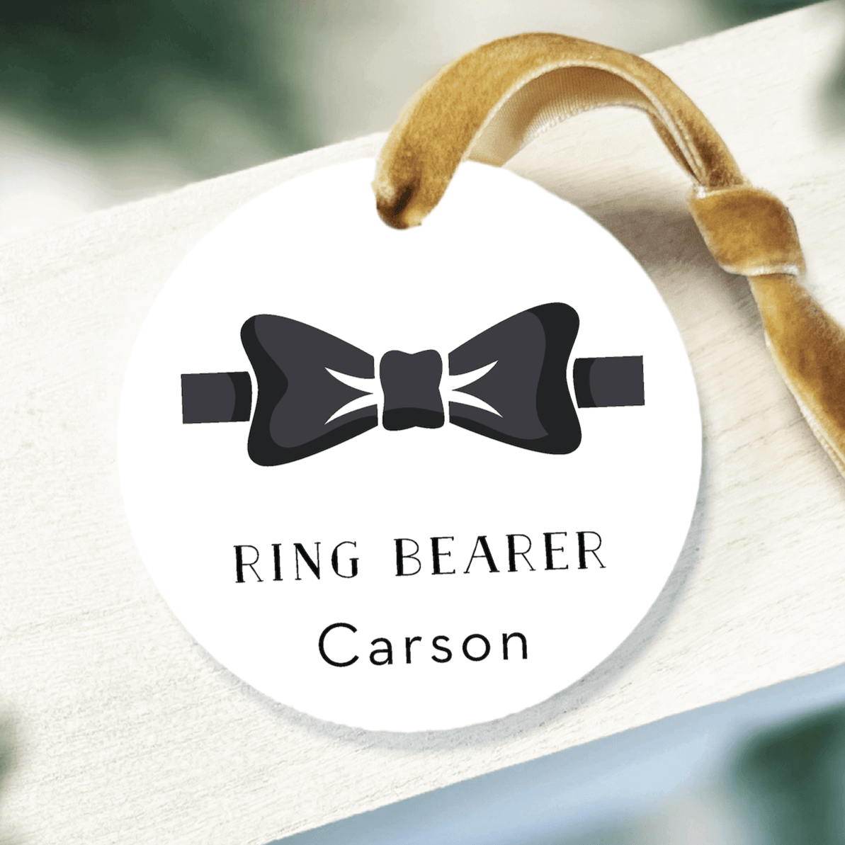 White round acrylic ornament with black bow tie icon, "RING BEARER" text, and personalized name "Carson" with tan ribbon loop.