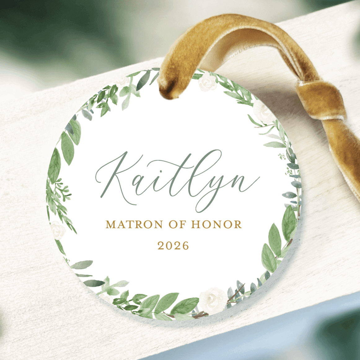 White acrylic ornament with green leafy wreath border, personalized name Kaitlyn, Matron of Honor 2026 text, and tan ribbon hanger.