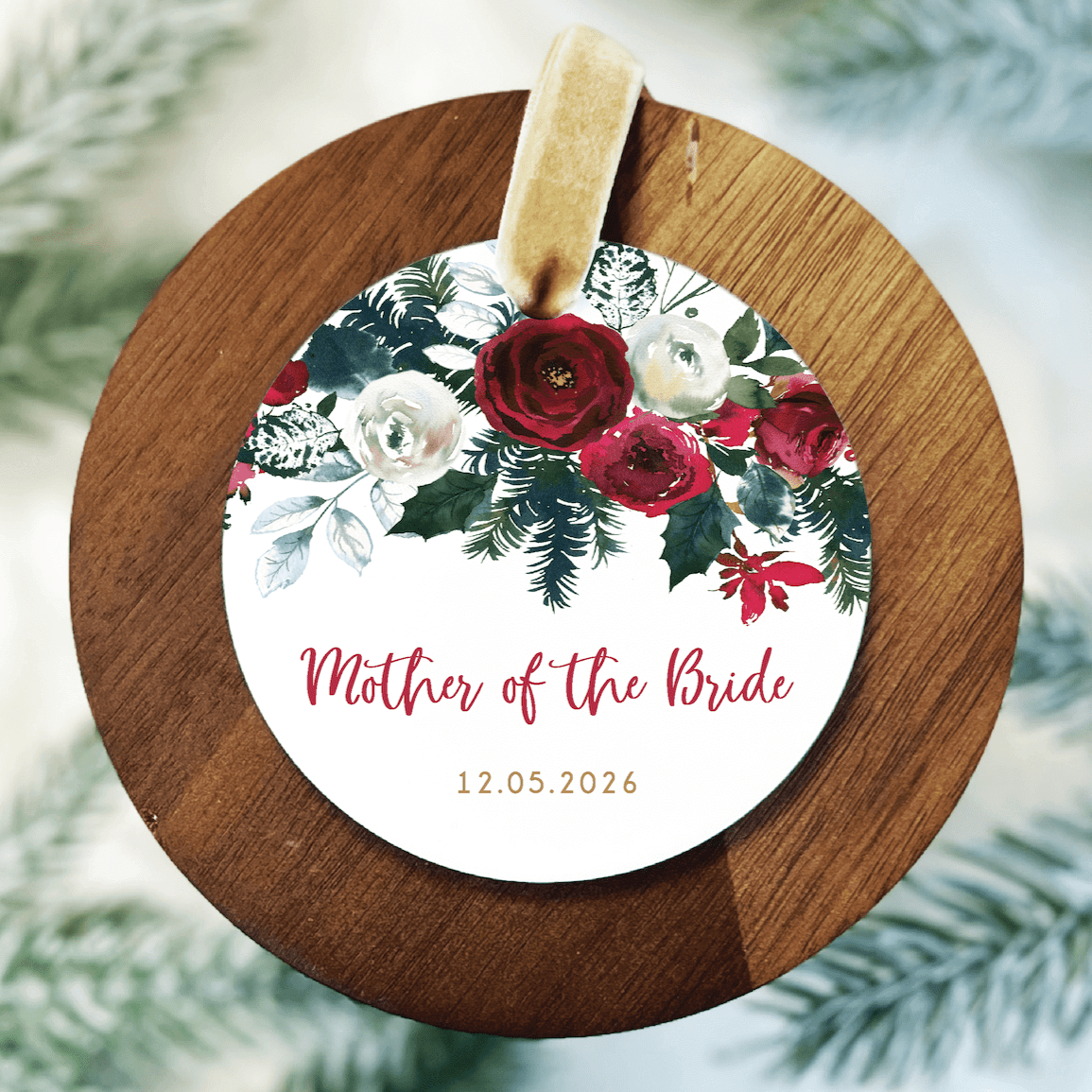 Wooden round ornament with white acrylic center featuring red and cream flowers, "Mother of the Bride" text, and date 12.05.2026, hung with tan ribbon.
