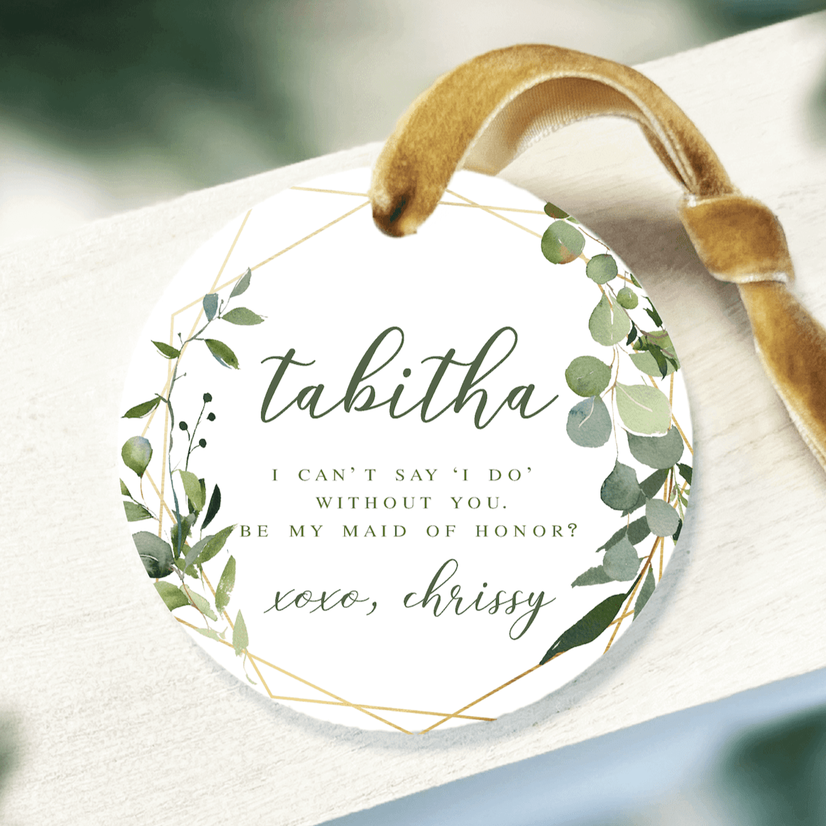 White acrylic ornament with greenery wreath design and personalized text asking "tabitha, i can't say 'i do' without you, be my maid of honor" signed "love, chrissy" with tan ribbon loop.