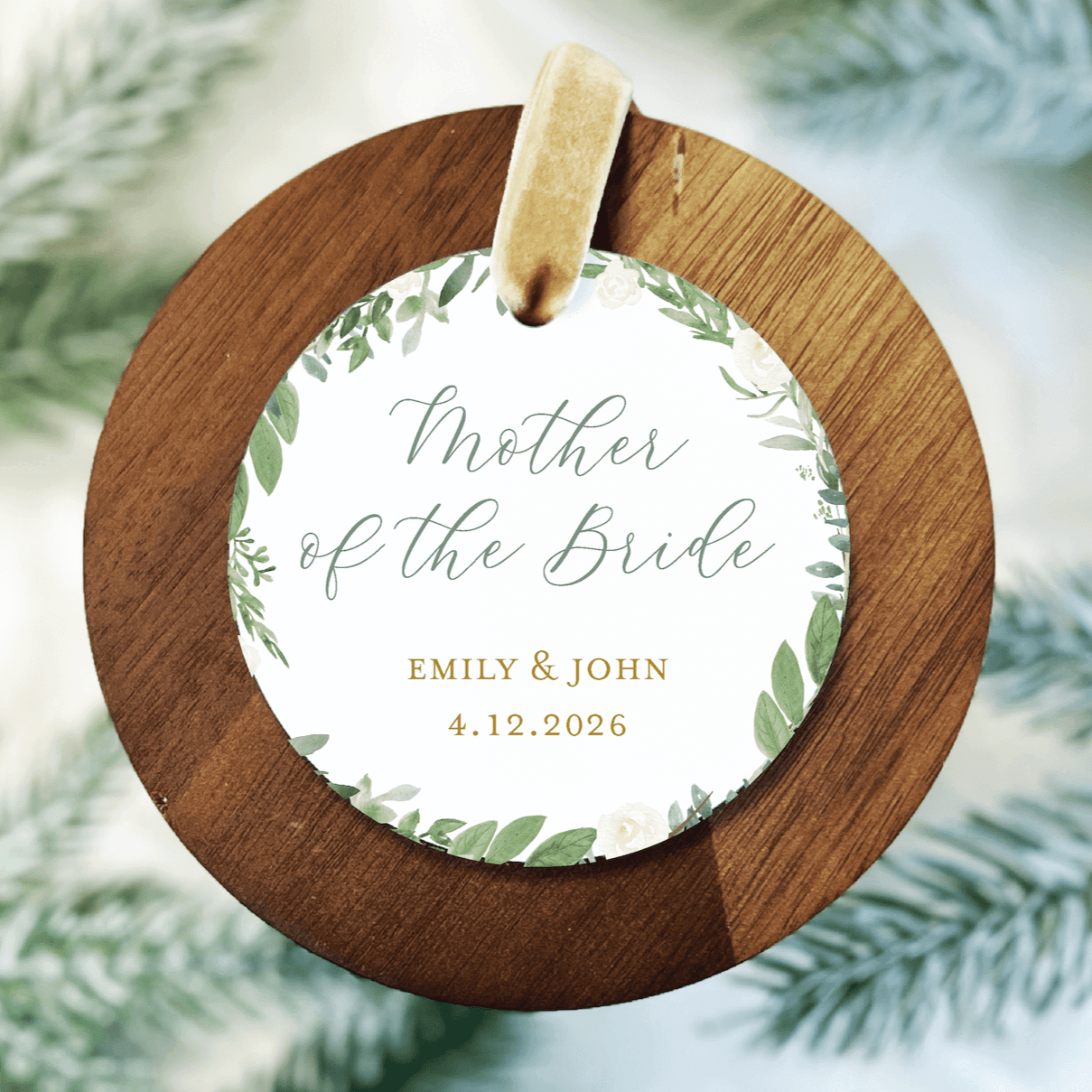 Round wooden ornament with white acrylic center featuring "Mother of the Bride" text, couple names Emily & John, and wedding date 4.12.2026 with green leaf design.
