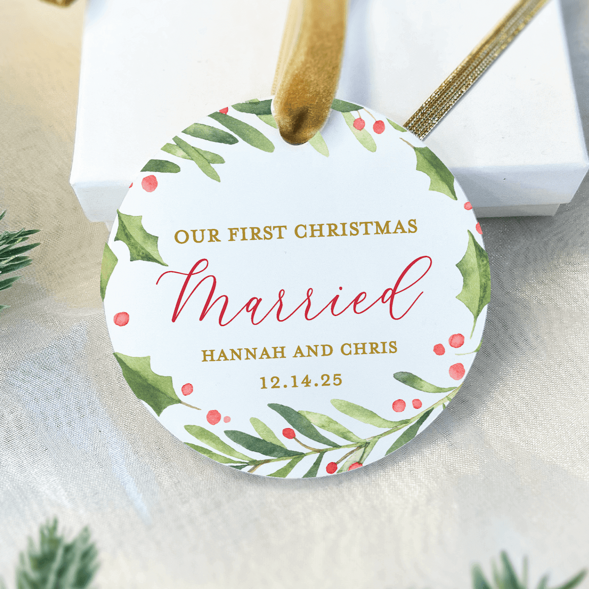 White acrylic ornament with holly greenery border, gold text reading "Our First Christmas Married" and personalized names "Hannah and Chris" with date 12.14.25, hung with twine.
