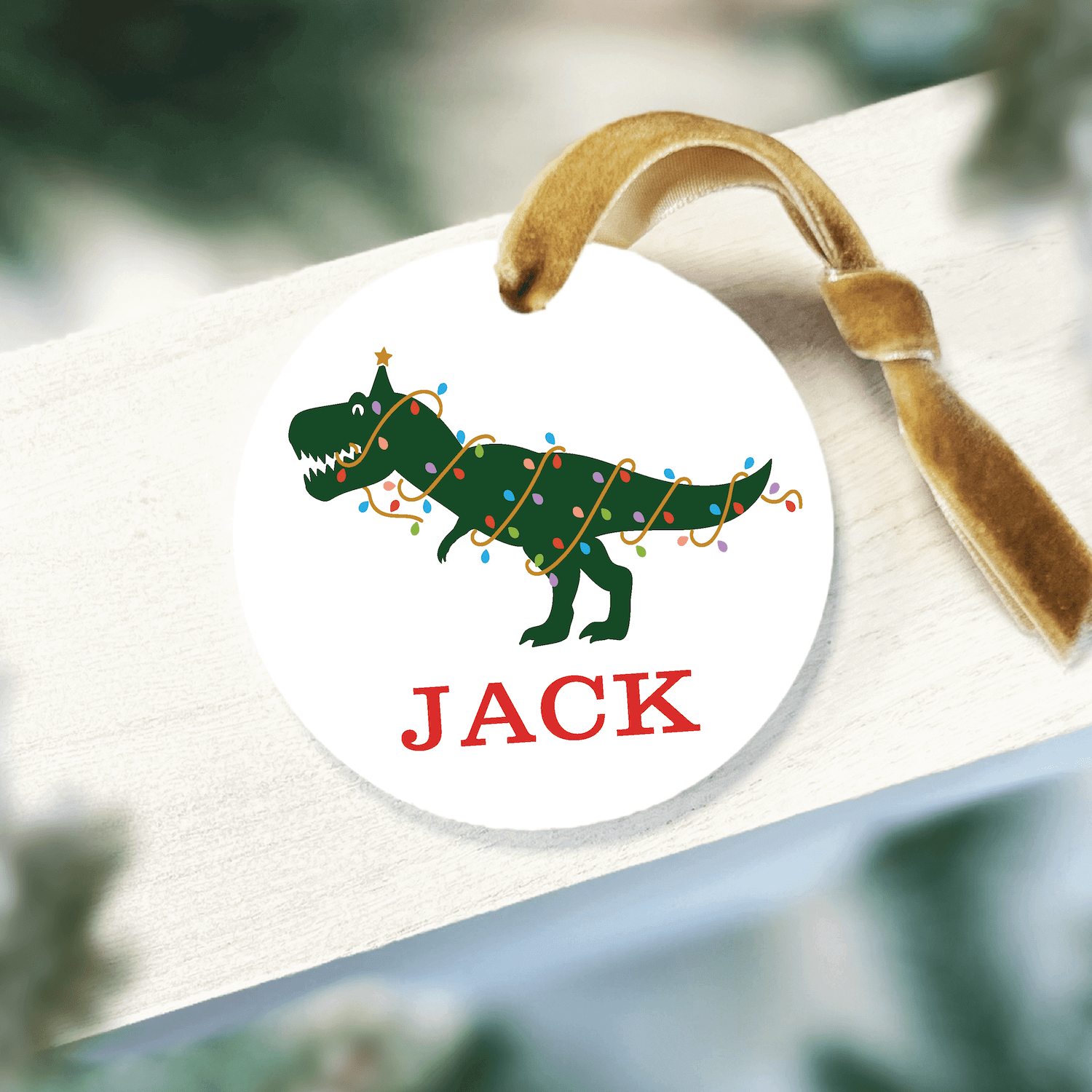 Round acrylic ornament with green T-Rex dinosaur wearing Christmas lights and red "JACK" text below, tan string attached.