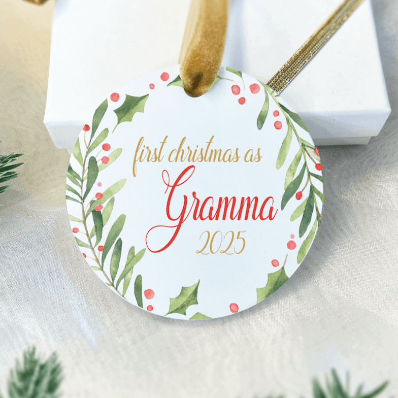 Round acrylic ornament with gold string, featuring "first christmas as Gramma" text in red and gold script surrounded by holly and berry wreath design.