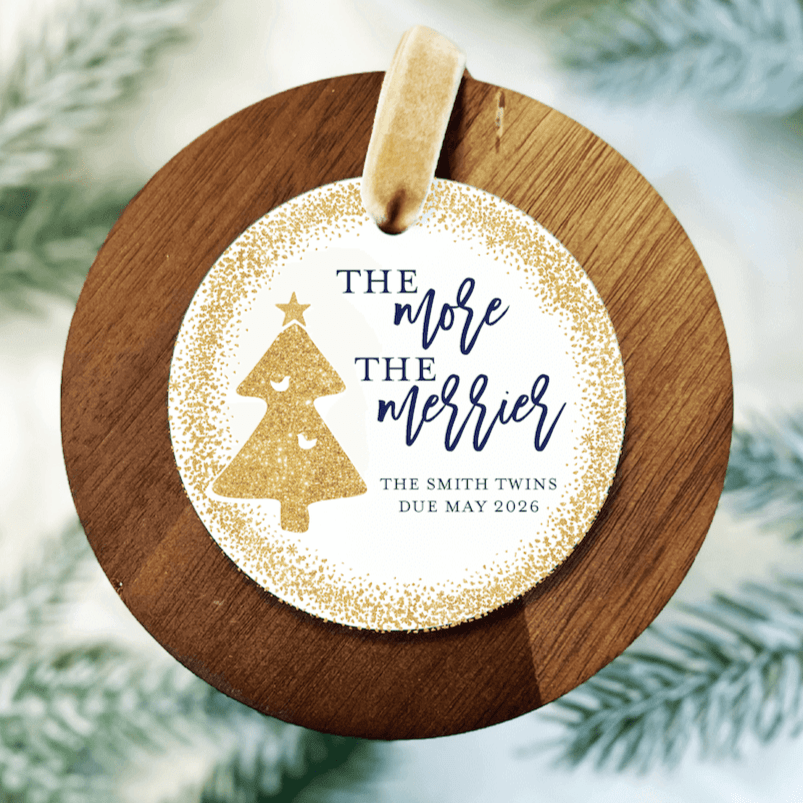 White acrylic ornament with gold glitter tree and "The more the merrier" text, personalized with names and due date, mounted on wooden backing with twine.