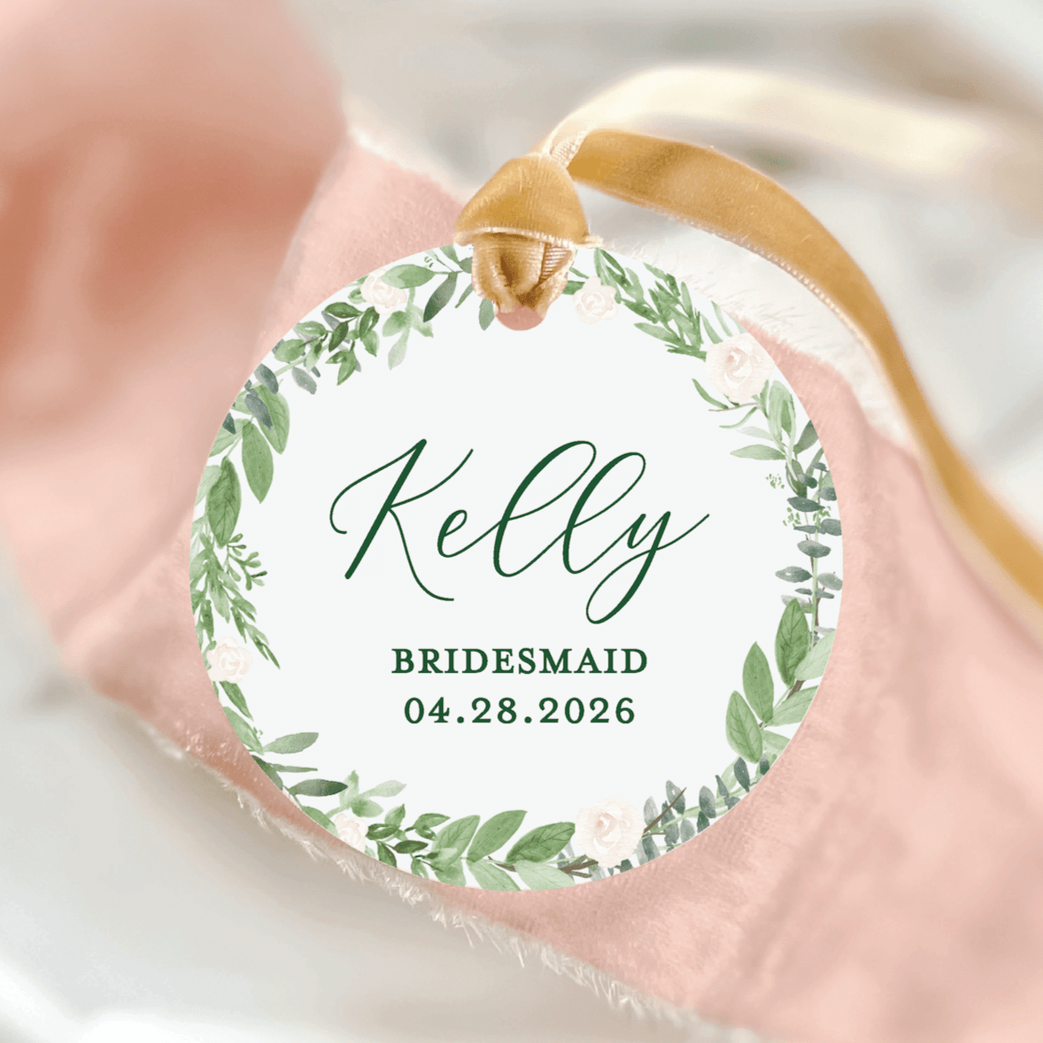 Round acrylic ornament with green leaf wreath design, personalized with "Kelly" and "BRIDESMAID 04.28.2026" text, gold ribbon attached.
