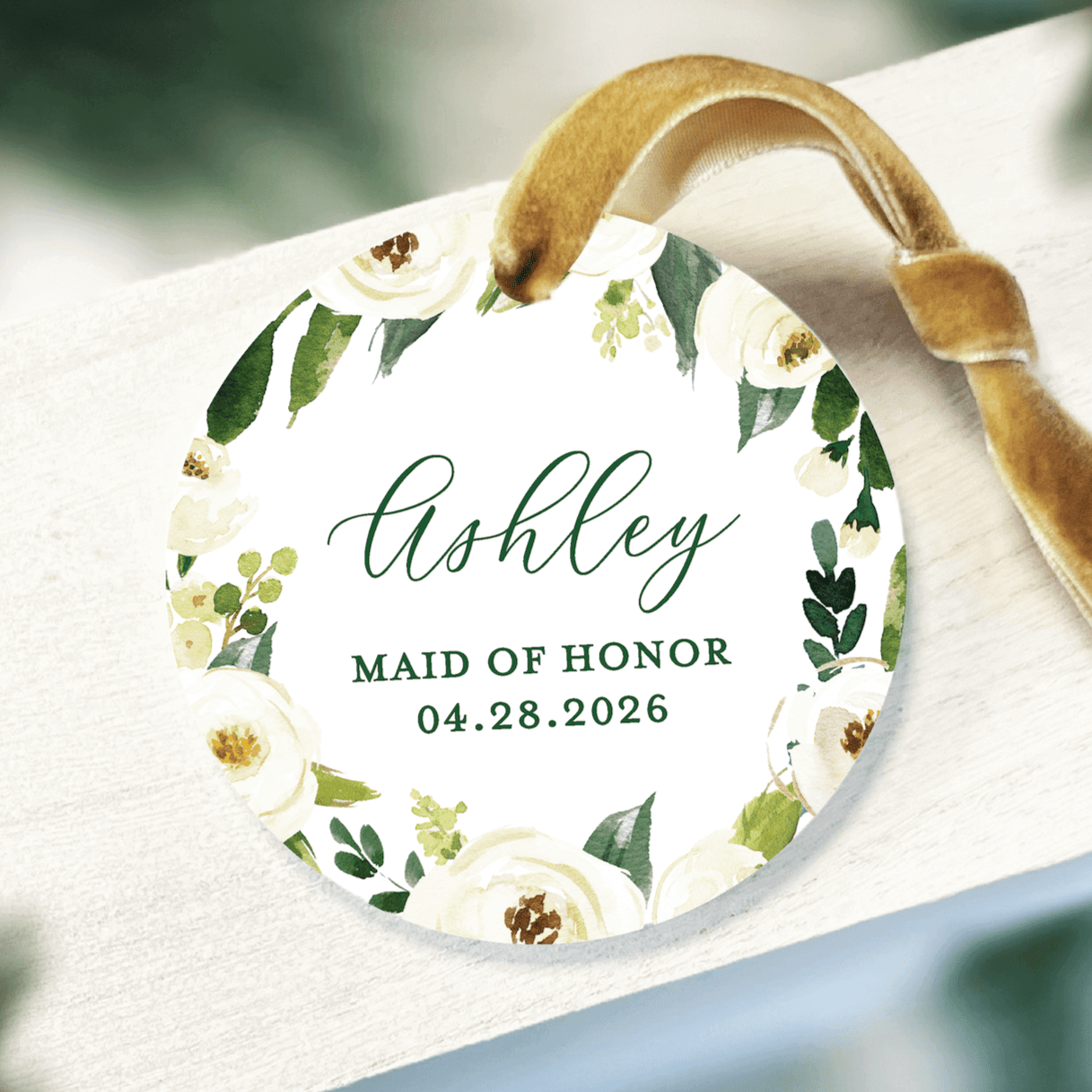 White acrylic ornament with gold floral border, personalized with name Ashley, Maid of Honor title, and wedding date 04.28.2026, attached with gold ribbon.
