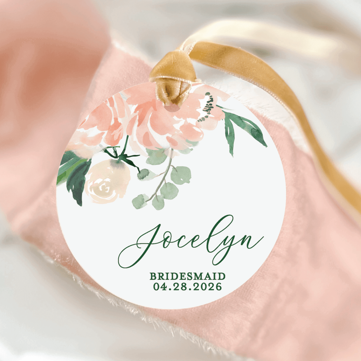 White acrylic ornament with pink and cream floral design, personalized with "Jocelyn" and "BRIDESMAID 04.28.2026" text, gold ribbon loop.