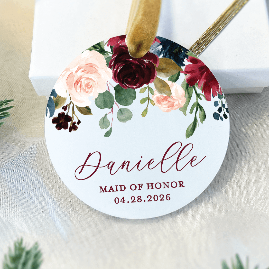 White acrylic ornament with floral design featuring pink and burgundy roses, personalized with "Danielle" and "MAID OF HONOR 04.28.2026" in burgundy script text, hanging from gold ribbon.