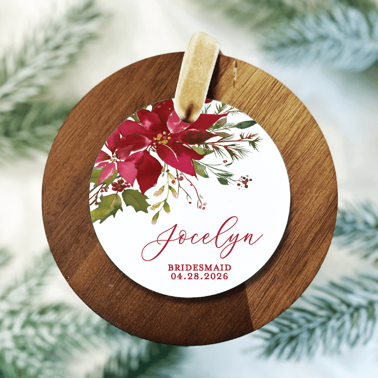 Wooden round ornament with white acrylic disc featuring red poinsettia design, personalized with "Jocelyn" and "BRIDESMAID 04.28.2026" text, gold ribbon loop.
