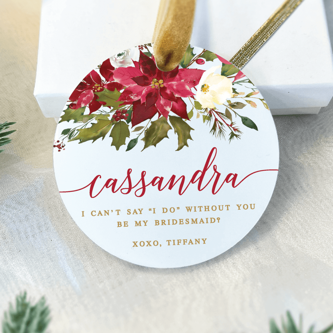 White acrylic ornament with burgundy and cream florals, personalized name "cassandra" in script, and bridesmaid proposal message in red and gold text with twine hanger.