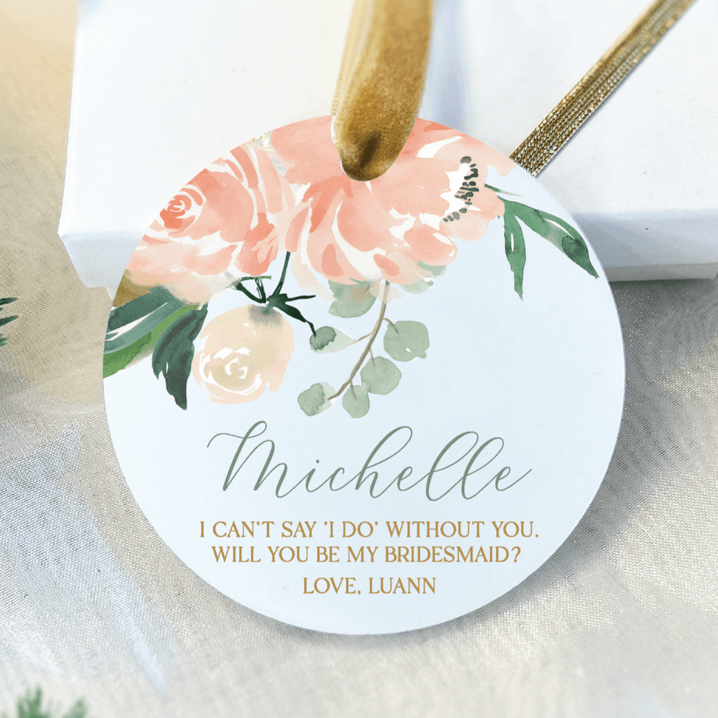 White round acrylic ornament with peach floral design, personalized name "Michelle" and bridesmaid proposal message in gold text with gold ribbon loop.