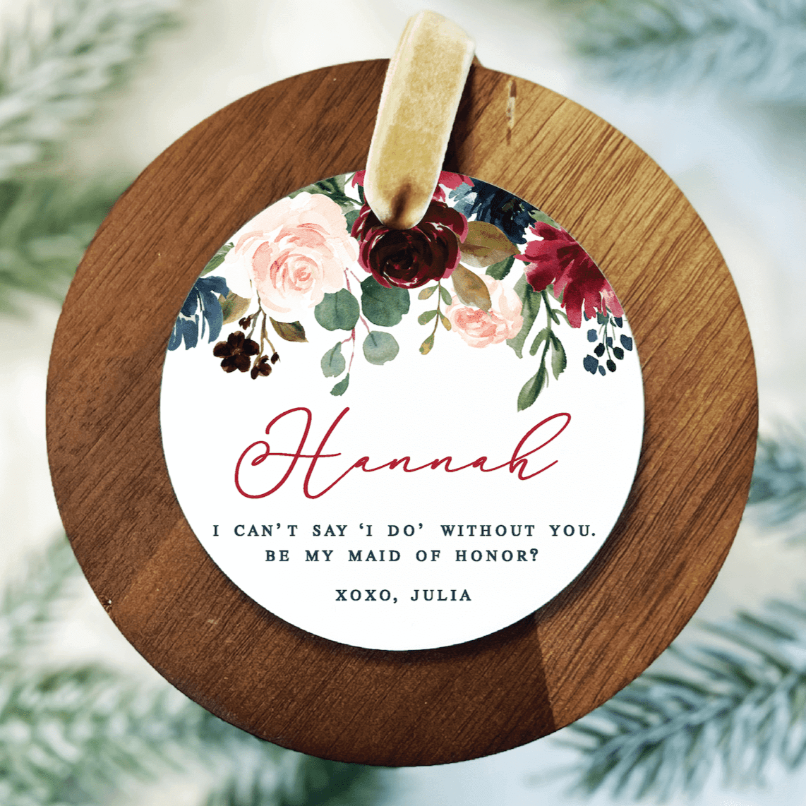 Round wooden ornament with white center featuring burgundy floral design, pink script text reading "Hannah," and personalized message asking someone to be maid of honor, signed "XOXO, JULIA"