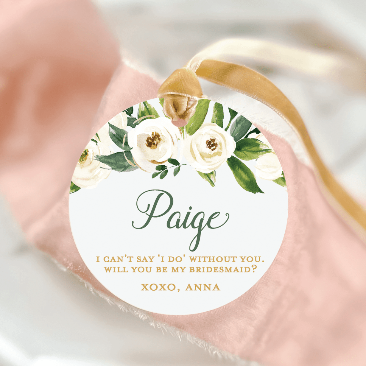 Round acrylic ornament with cream and gold floral design, personalized text reading "Paige" and bridesmaid proposal message, attached with gold ribbon.