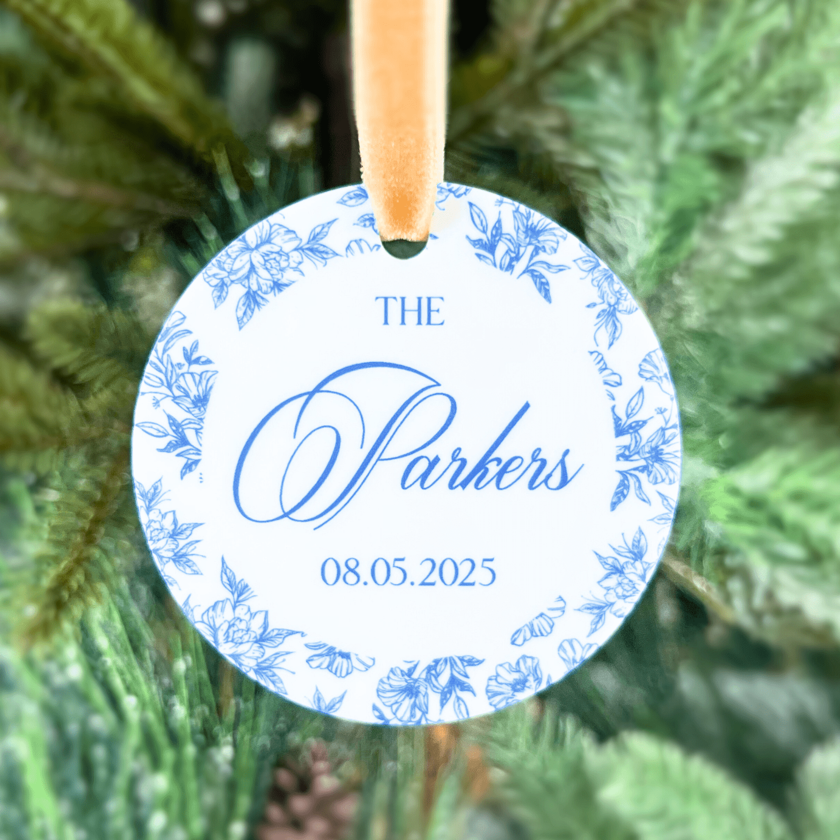 Round acrylic ornament with blue toile floral border, personalized with "The Parkers" script text and date 08.05.2025, hanging on evergreen branch.
