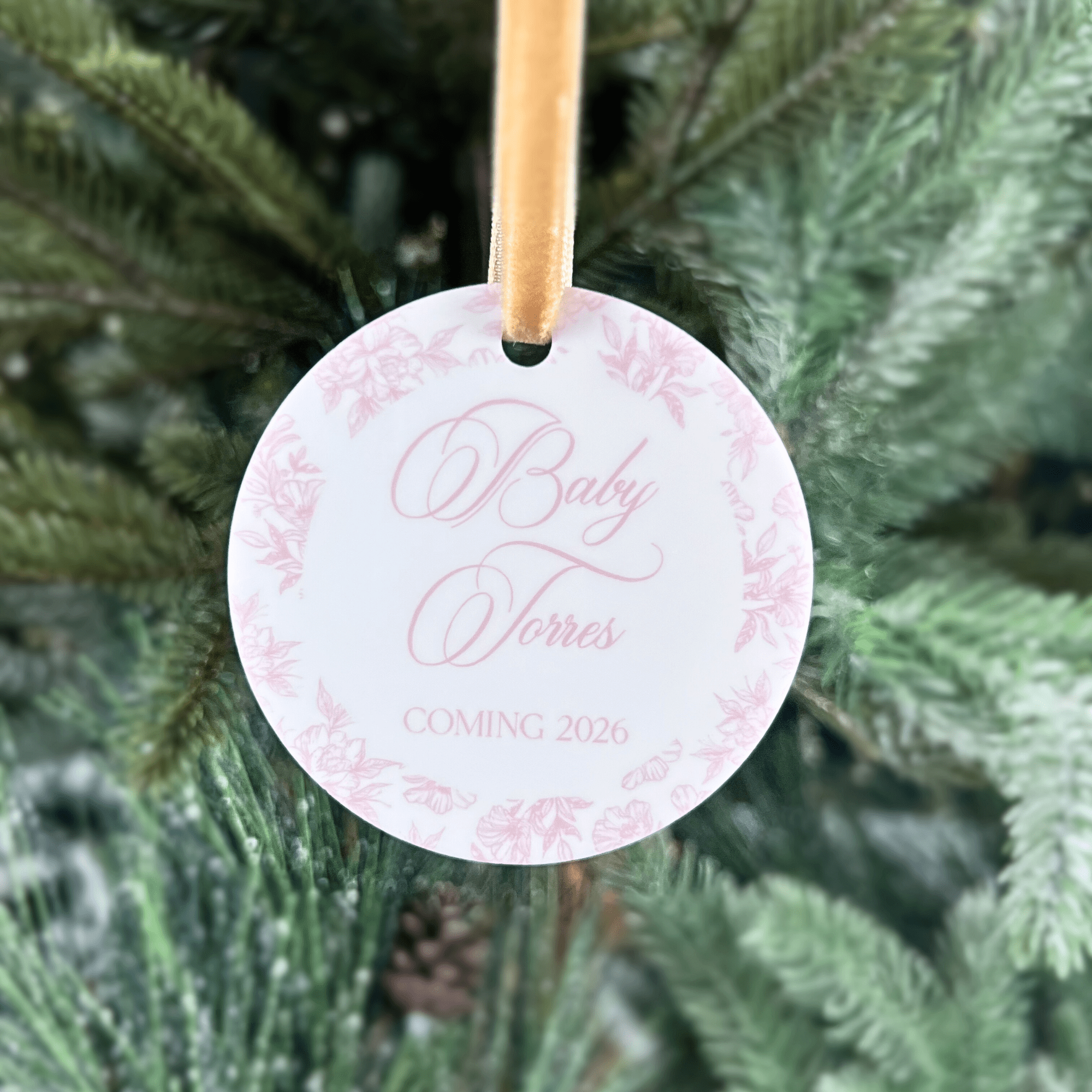 Pink floral toile ornament with "Baby James Coming 2026" text, hanging on evergreen branch with tan ribbon.