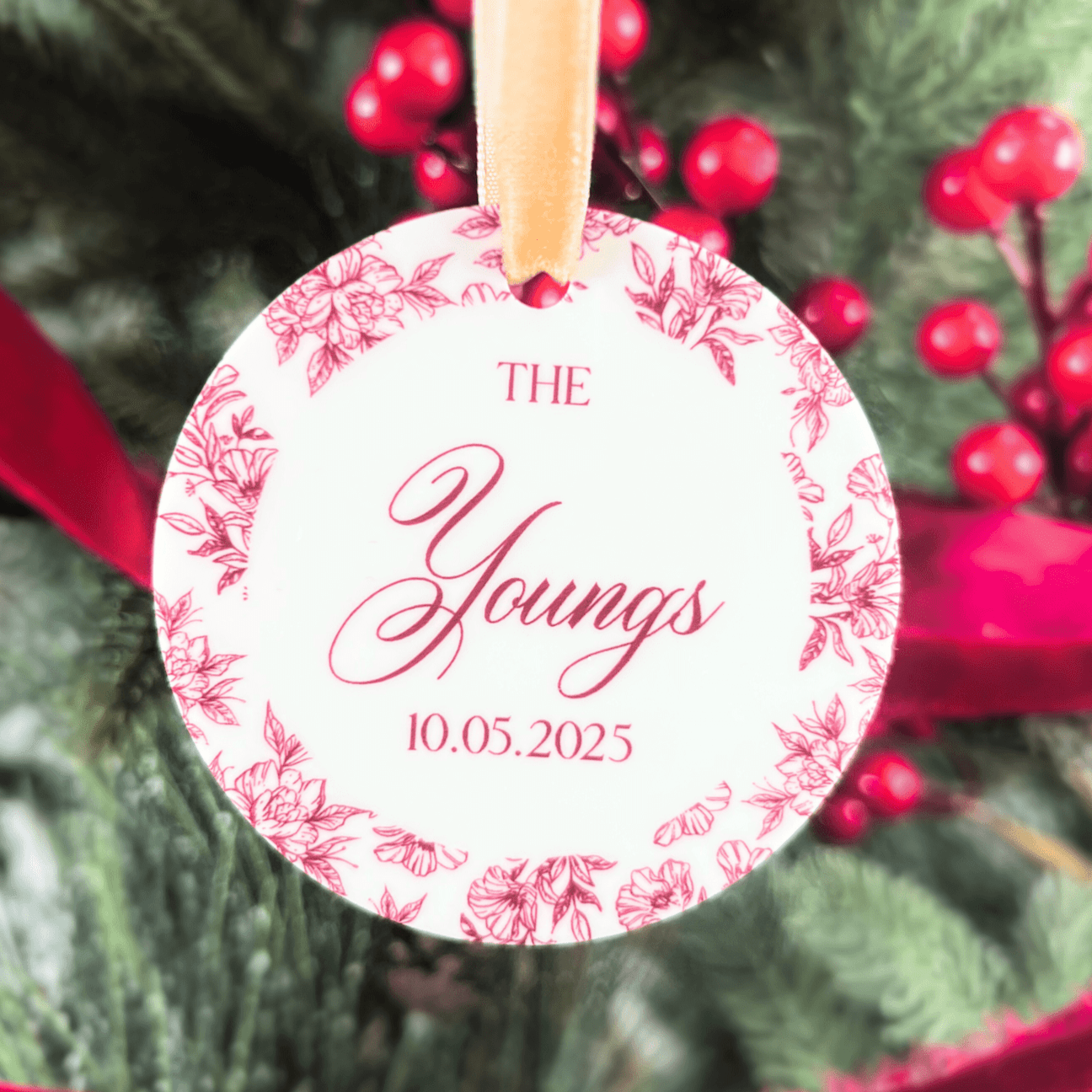 White acrylic ornament with pink toile floral border featuring "The Youngs" and date "10.05.2025" in script lettering, displayed on evergreen branches with red berries.
