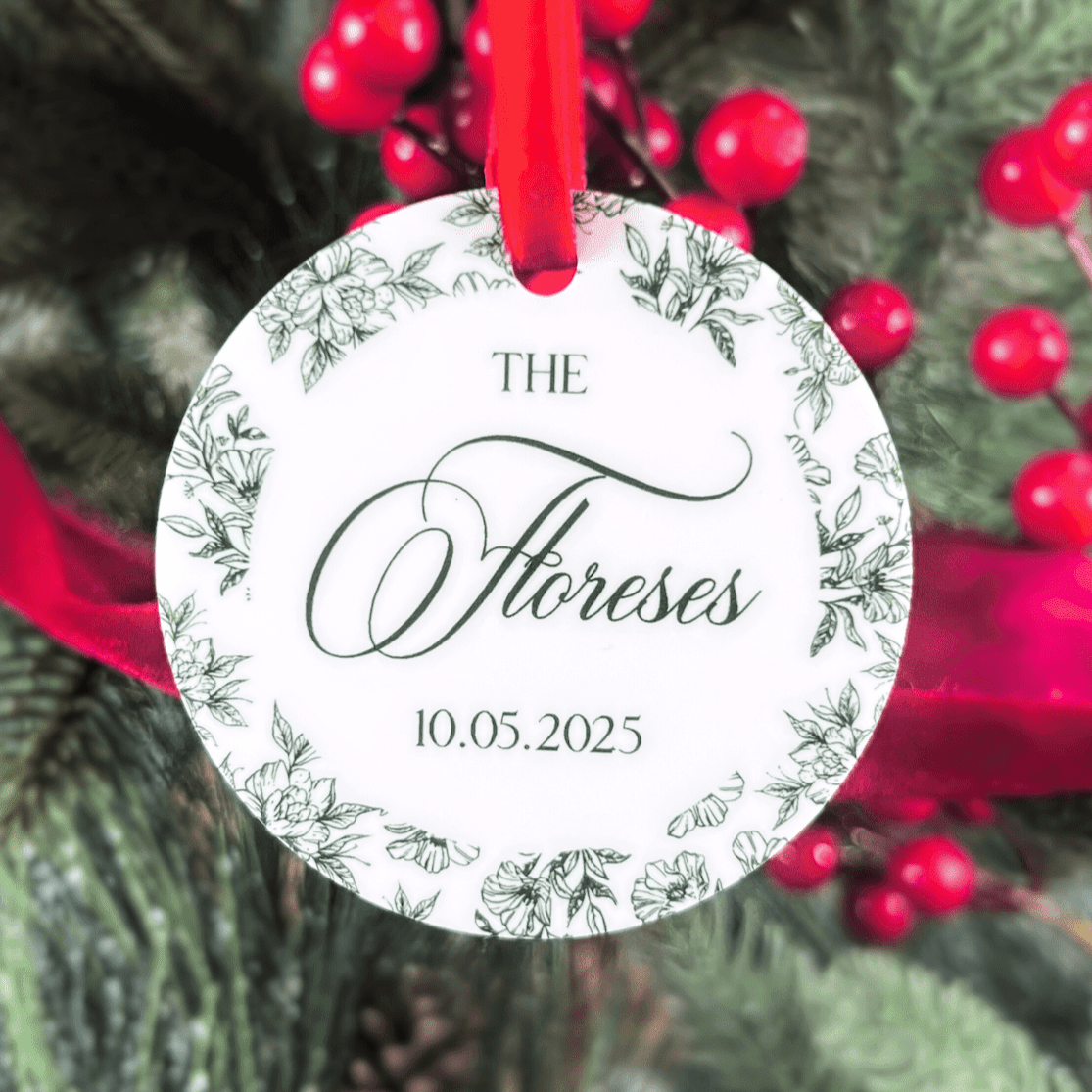 Round acrylic ornament with red ribbon, featuring "The Floreses" in script and date 10.05.2025 surrounded by delicate botanical line drawings.