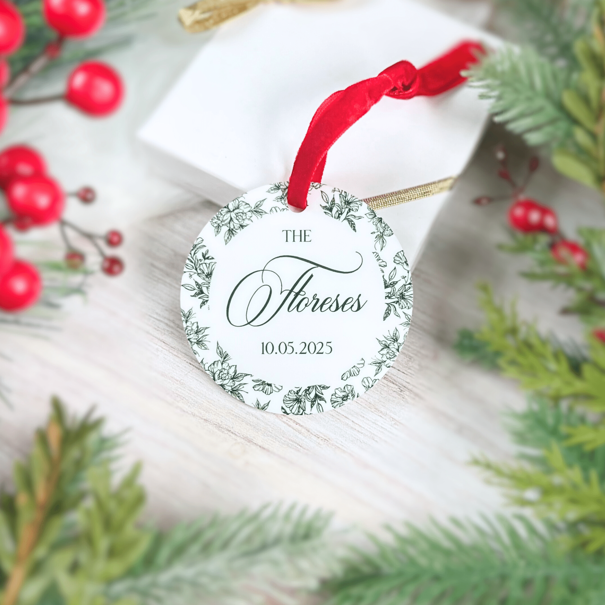 Round acrylic ornament with red ribbon featuring "The Moroses" and date 10.05.2025 in wreath design, surrounded by evergreen branches and red berries.