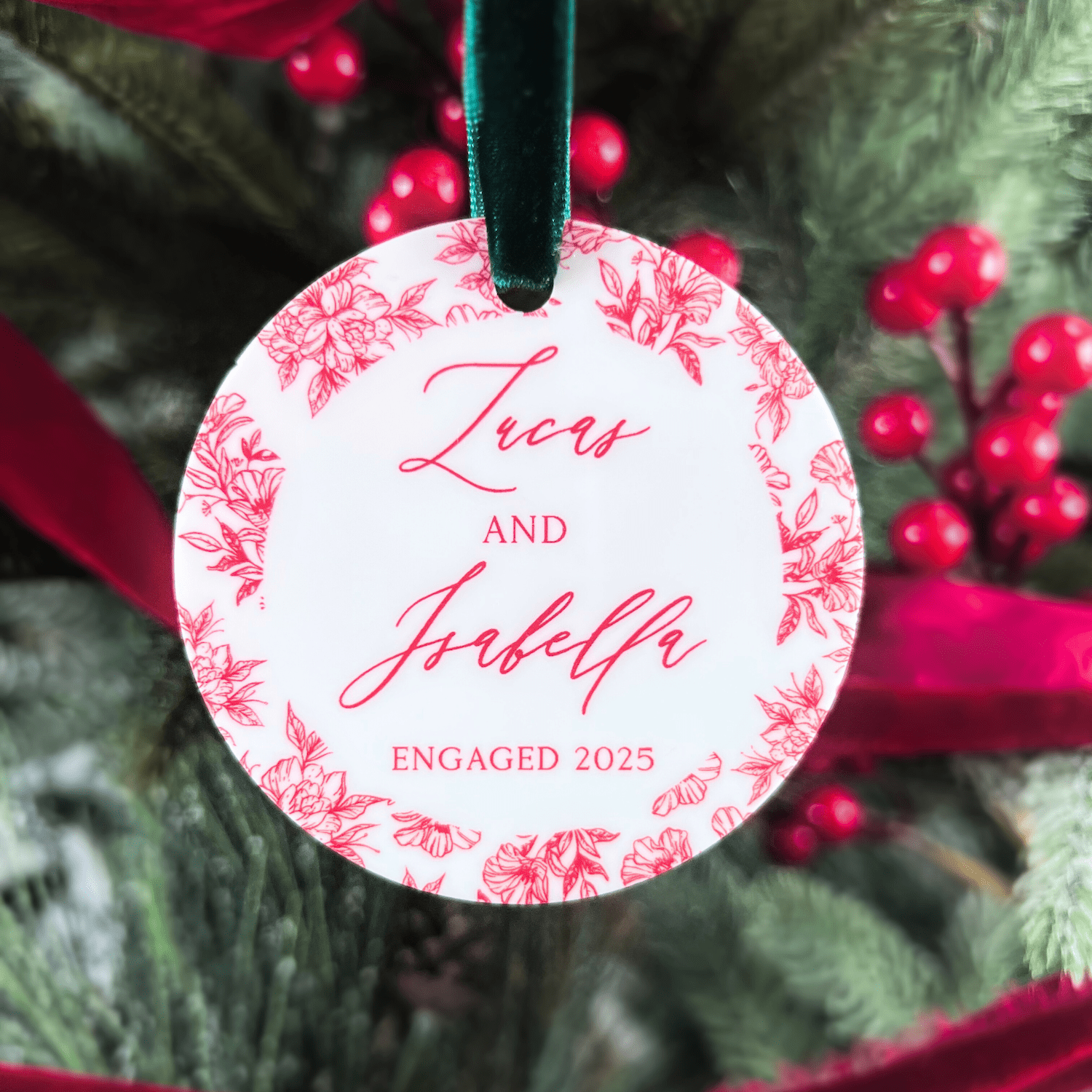 Round acrylic ornament with pink toile floral border and personalized names "Zoey and Jedediah" with "Engaged 2025" text, hanging from teal ribbon on evergreen branch with berries.