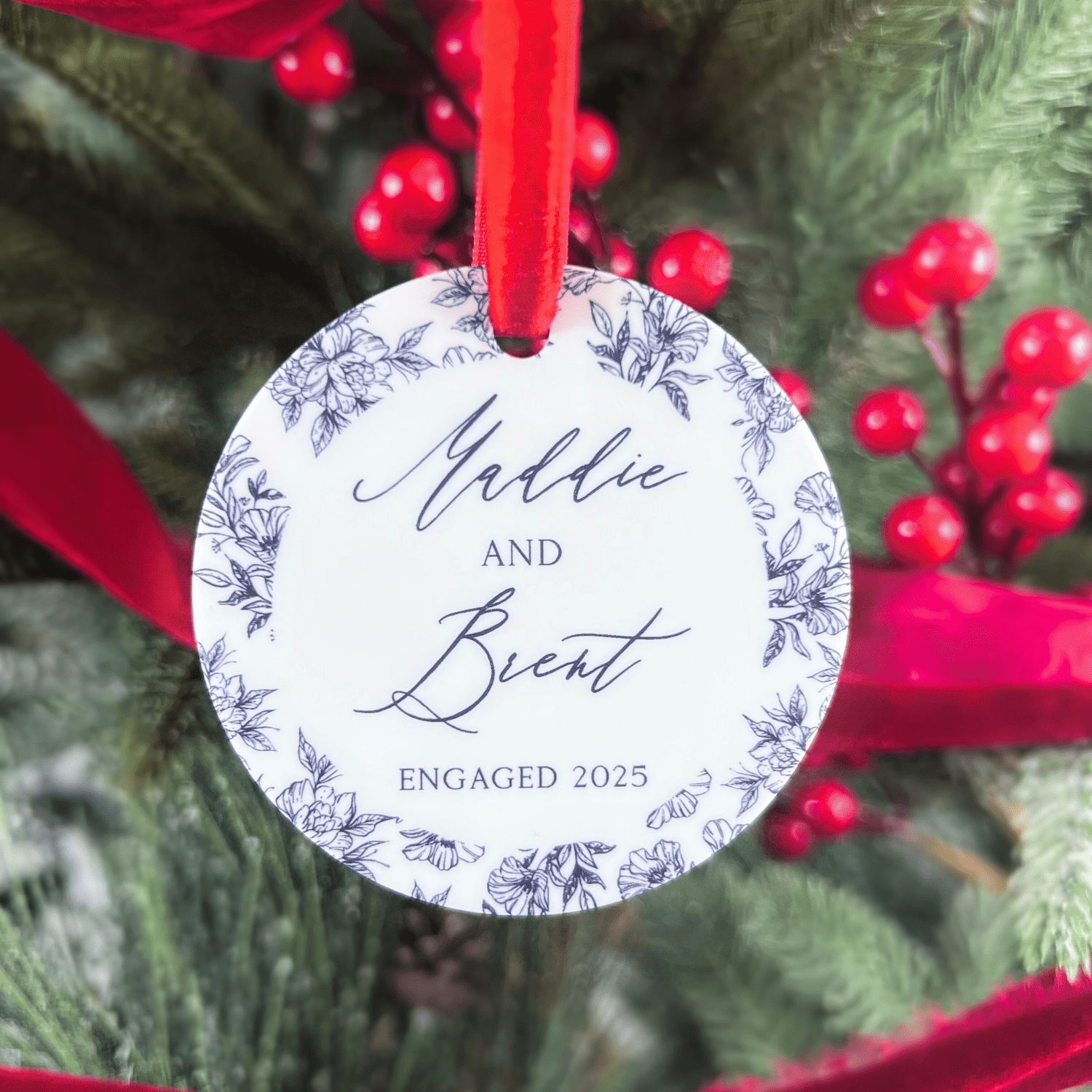 Round acrylic ornament with red ribbon, featuring blue toile floral border and personalized text reading "Addie and Brent ENGAGED 2025" hanging on evergreen branch with red berries.