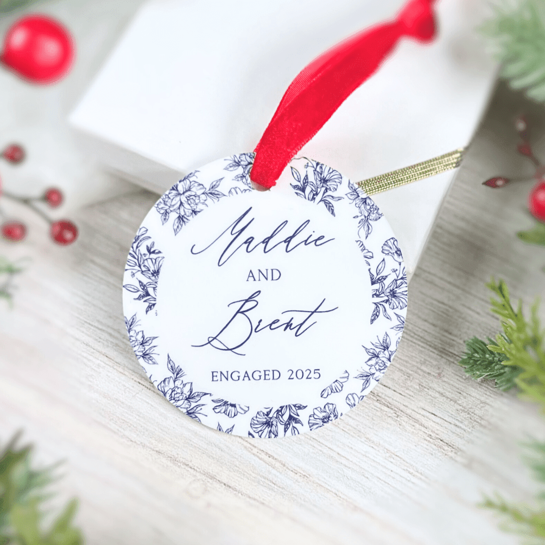 White acrylic ornament with blue toile floral border and personalized text reading "Millie and Brent Engaged 2025" tied with red ribbon.