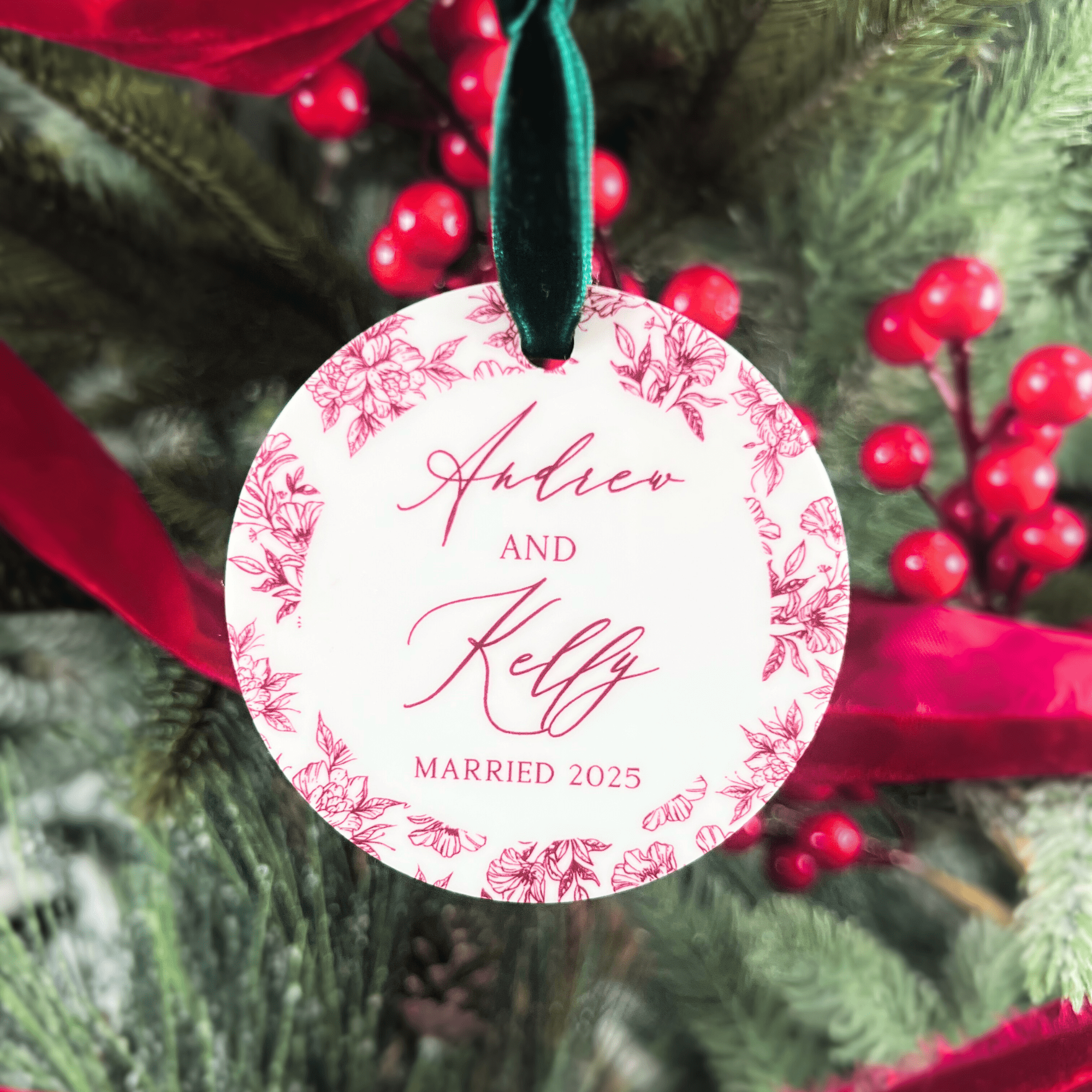 Round acrylic ornament with pink toile floral border featuring personalized names "Ashton and Kelly" and "Married 2025" text, hanging from green ribbon on evergreen branches.