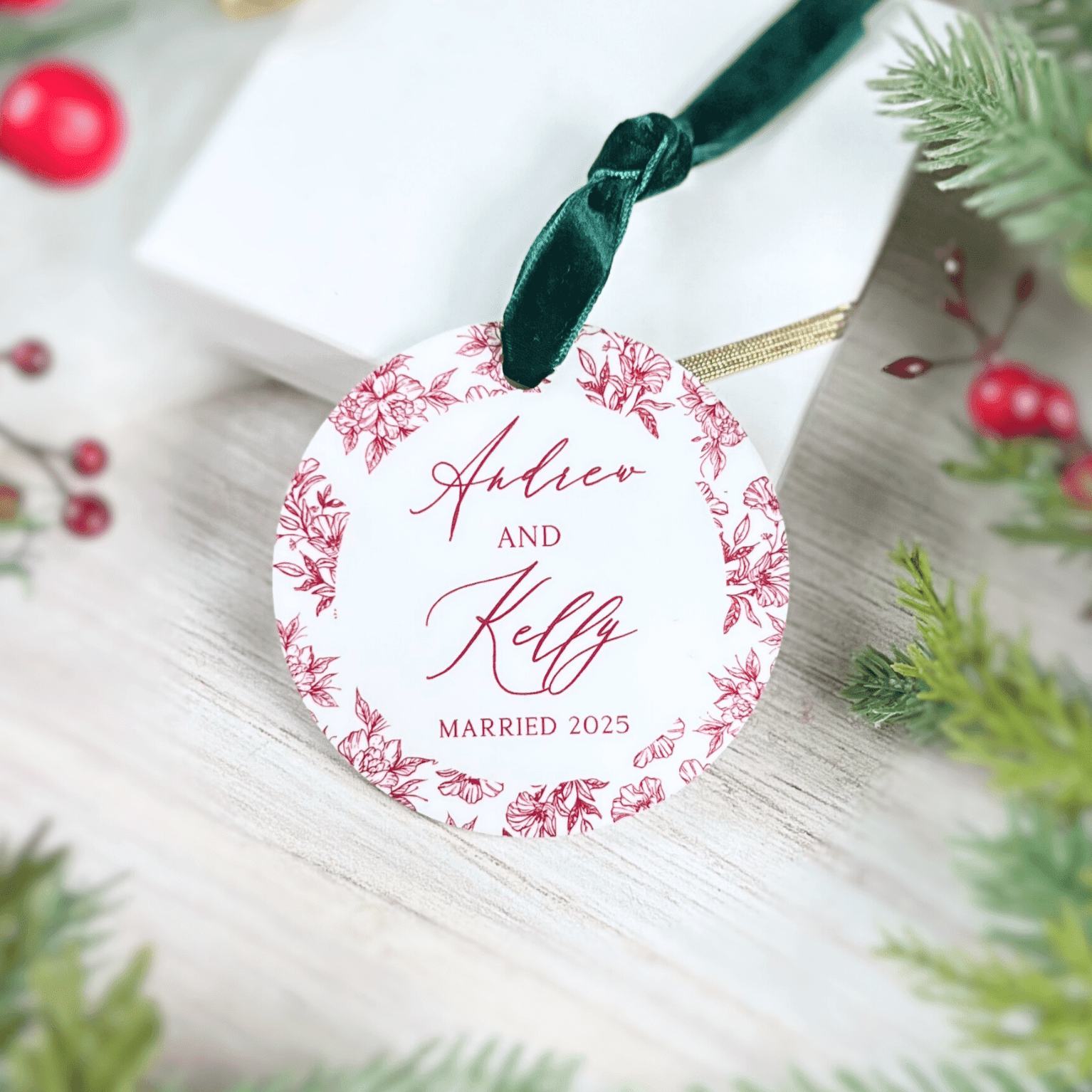 Round acrylic ornament with pink floral border and personalized names Ashton and Kelly with "married 2025" text, hung with dark green ribbon.