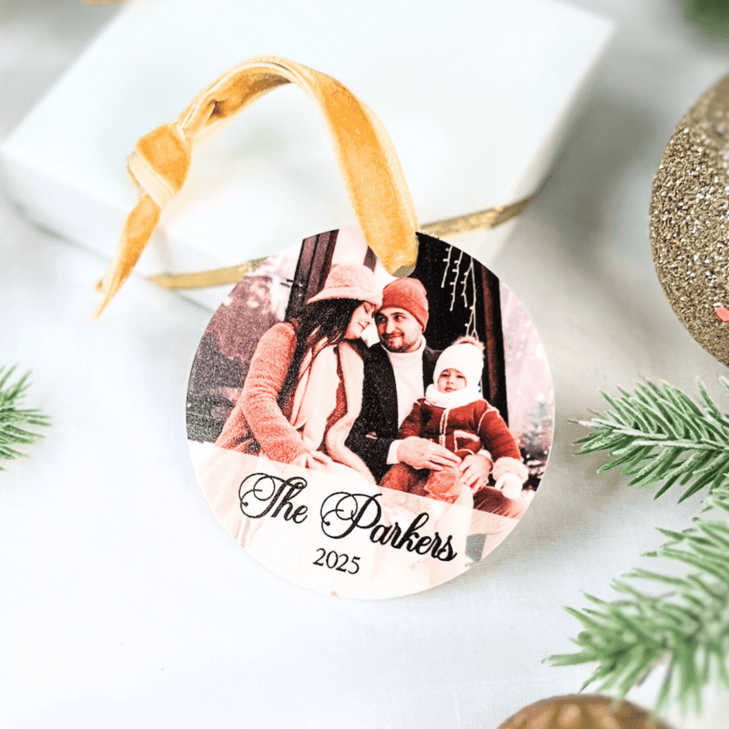 Oval acrylic ornament with family photo and "The Parkers 2023" text, gold ribbon attached, displayed on white surface with evergreen sprigs.