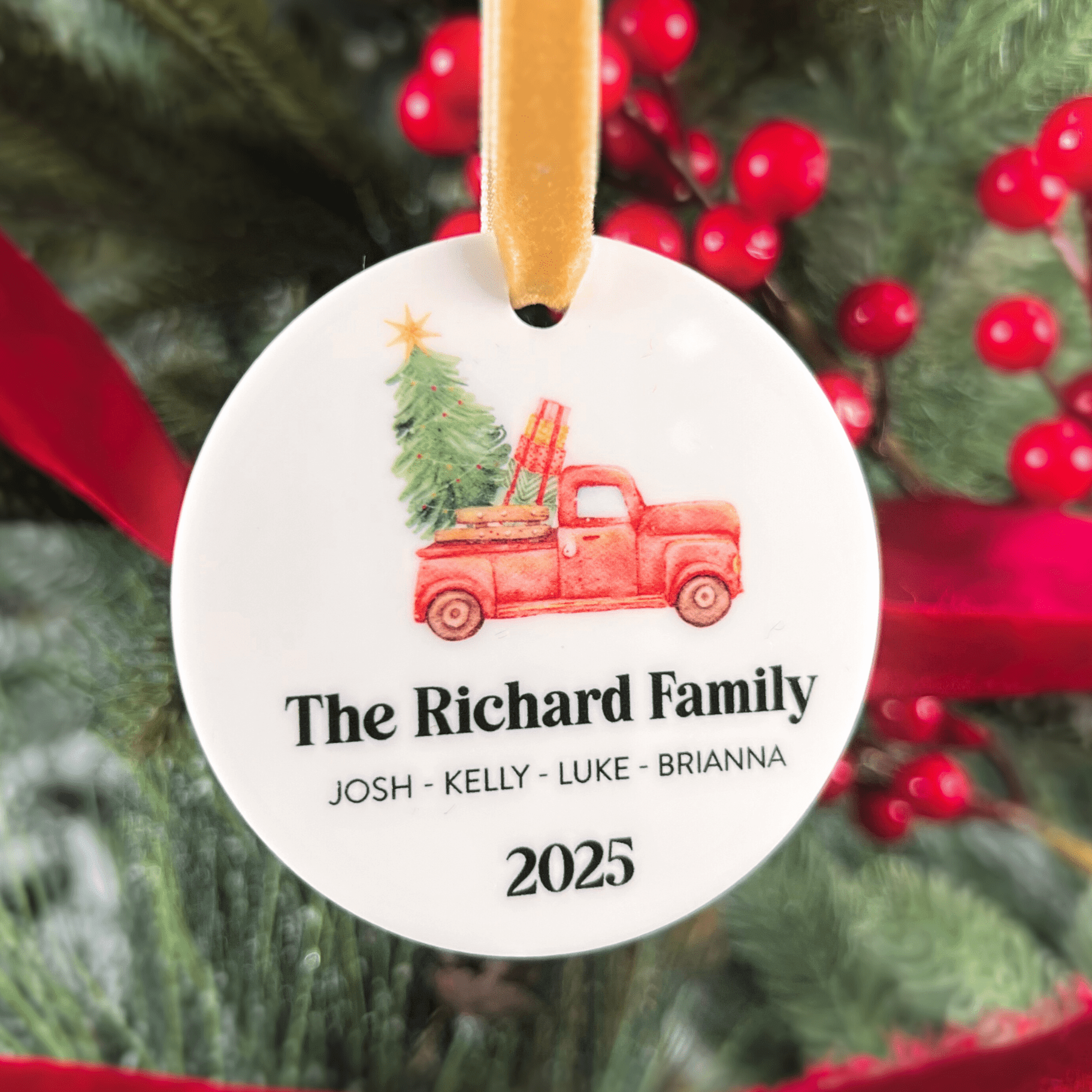 Round white acrylic ornament with red truck carrying Christmas tree design, family name and personalized names with 2025 date, gold ribbon.