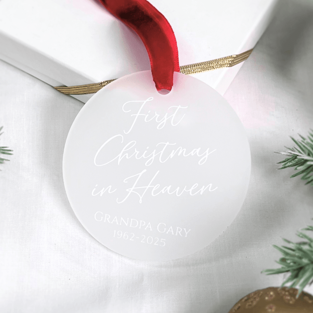 White acrylic circular ornament with red ribbon and gold accents, engraved with "First Christmas in Heaven" and personalized name and date.