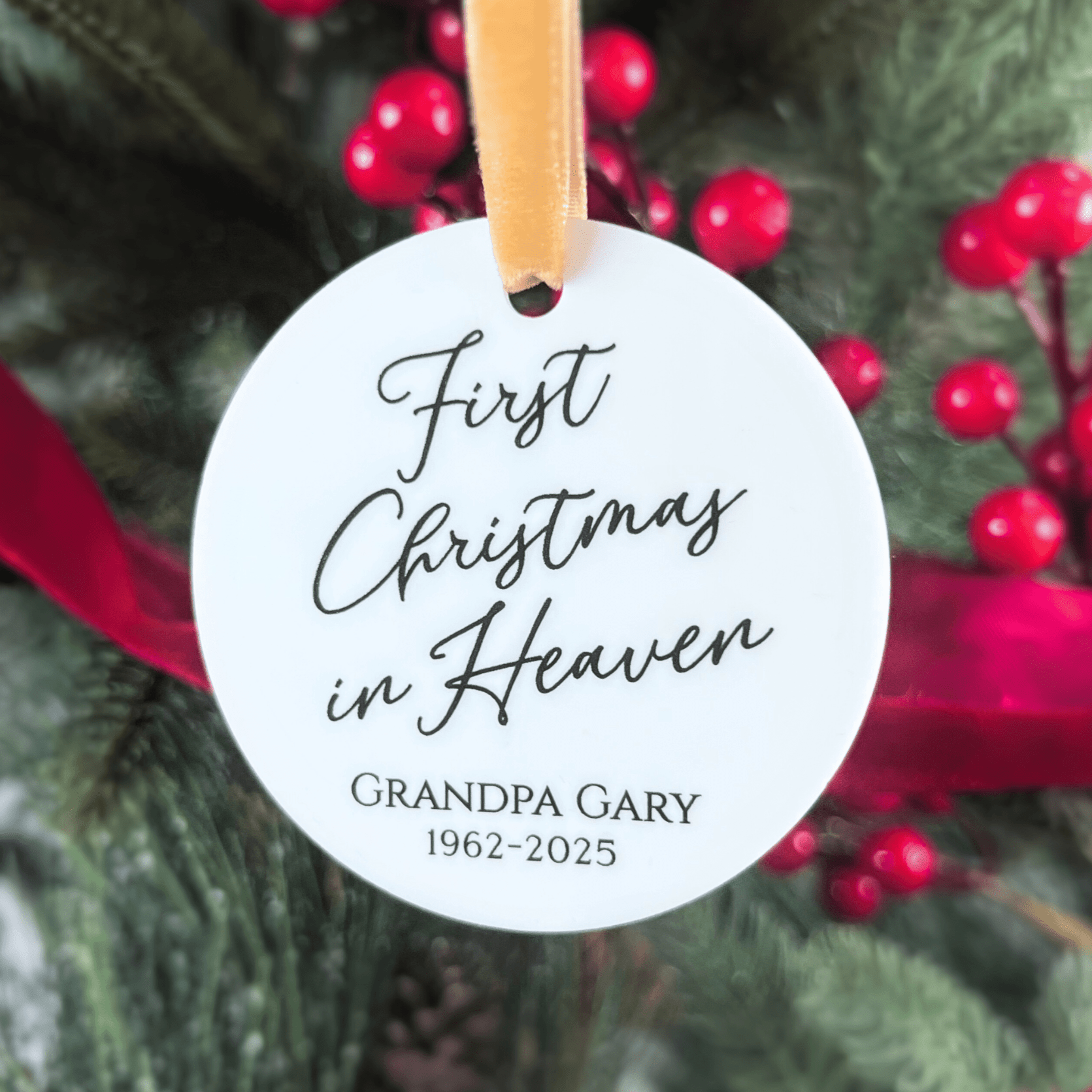 White round acrylic ornament with tan ribbon, personalized with "First Christmas in Heaven" and "GRANDPA GARY 1962-2025" in script text, hanging on evergreen branch with red berries.