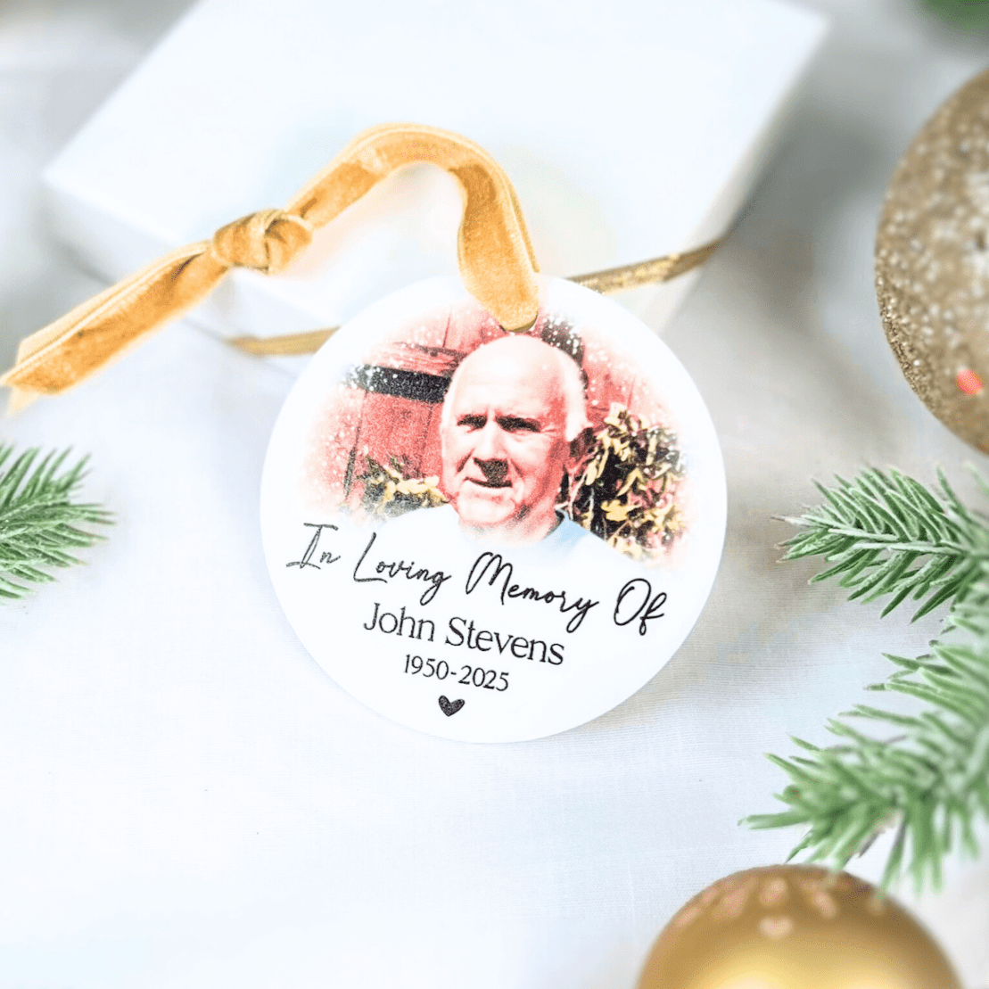 White acrylic memorial ornament with photo, "In Loving Memory Of" text, name and dates, gold ribbon, surrounded by evergreen sprigs and gold ornaments.