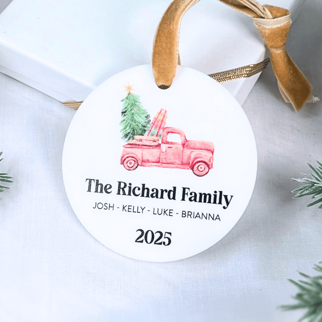 White round acrylic ornament with pink truck carrying Christmas tree, family name and four member names, gold ribbon hanger, 2025 date
