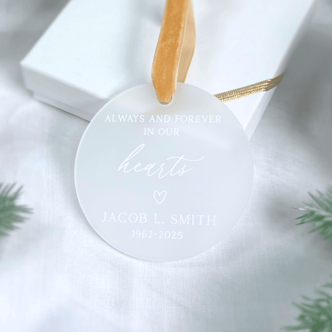 White acrylic memorial ornament with gold ribbon, engraved with "Always and Forever in Our Hearts," a heart icon, and personalized name and dates.