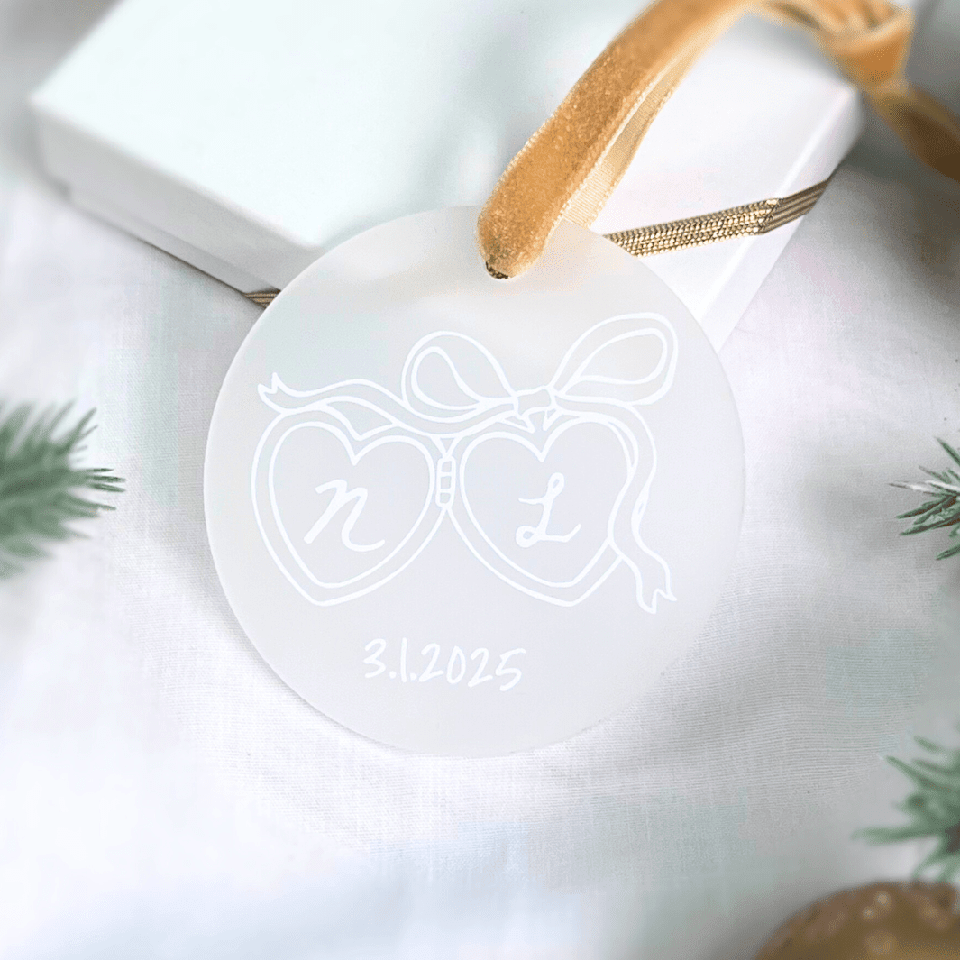 Frosted acrylic ornament with two linked hearts engraved with initials "n" and "l" and date "31.2025" below, attached with gold ribbon.