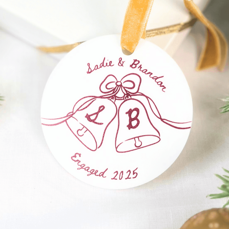 White acrylic ornament with pink line art of two wedding bells tied with a bow, personalized with initials L and B, names Jodie and Brandon, and Engaged 2025 text.