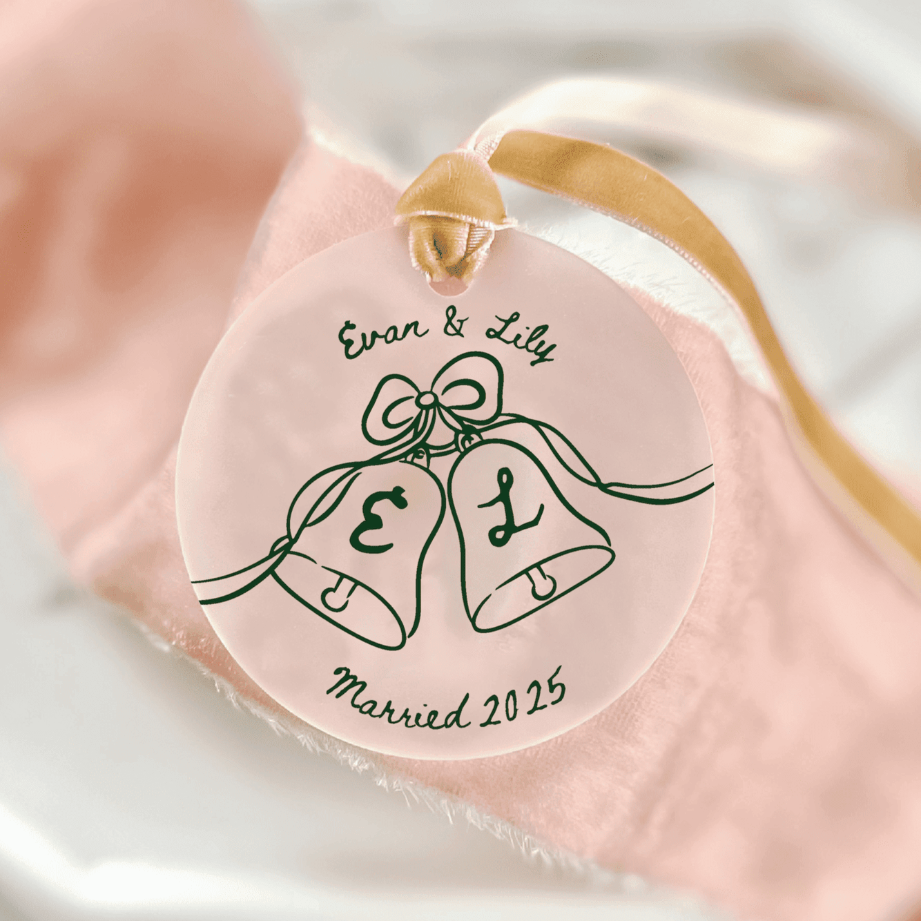 Pink acrylic ornament with gold ribbon featuring two bells, initials E and L, and "Evan & Lib Married 2025" text.